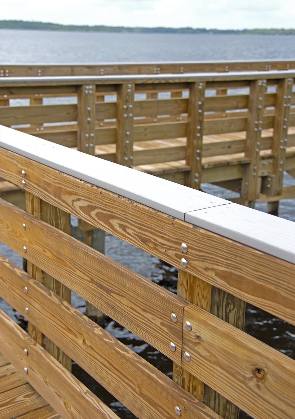 Close-up of a wooden dock with horizontal wooden planks and metal bolts, extending over water with an open water view and cloudy sky in the background.