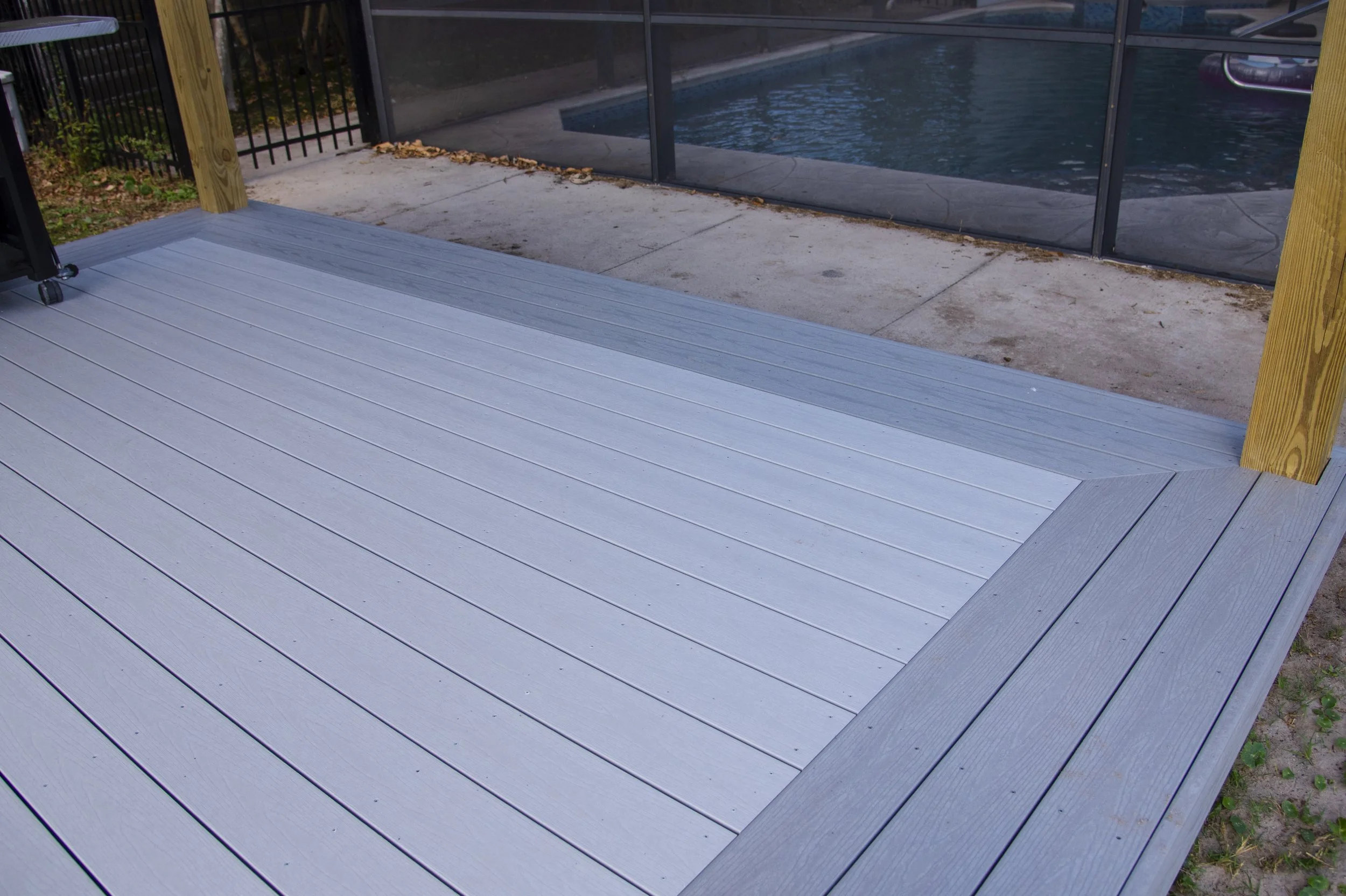View of a backyard patio with gray decking and an adjacent in-ground swimming pool enclosed by a glass screen. Custom decking, composite material. Built by Orlando expert deck builder.