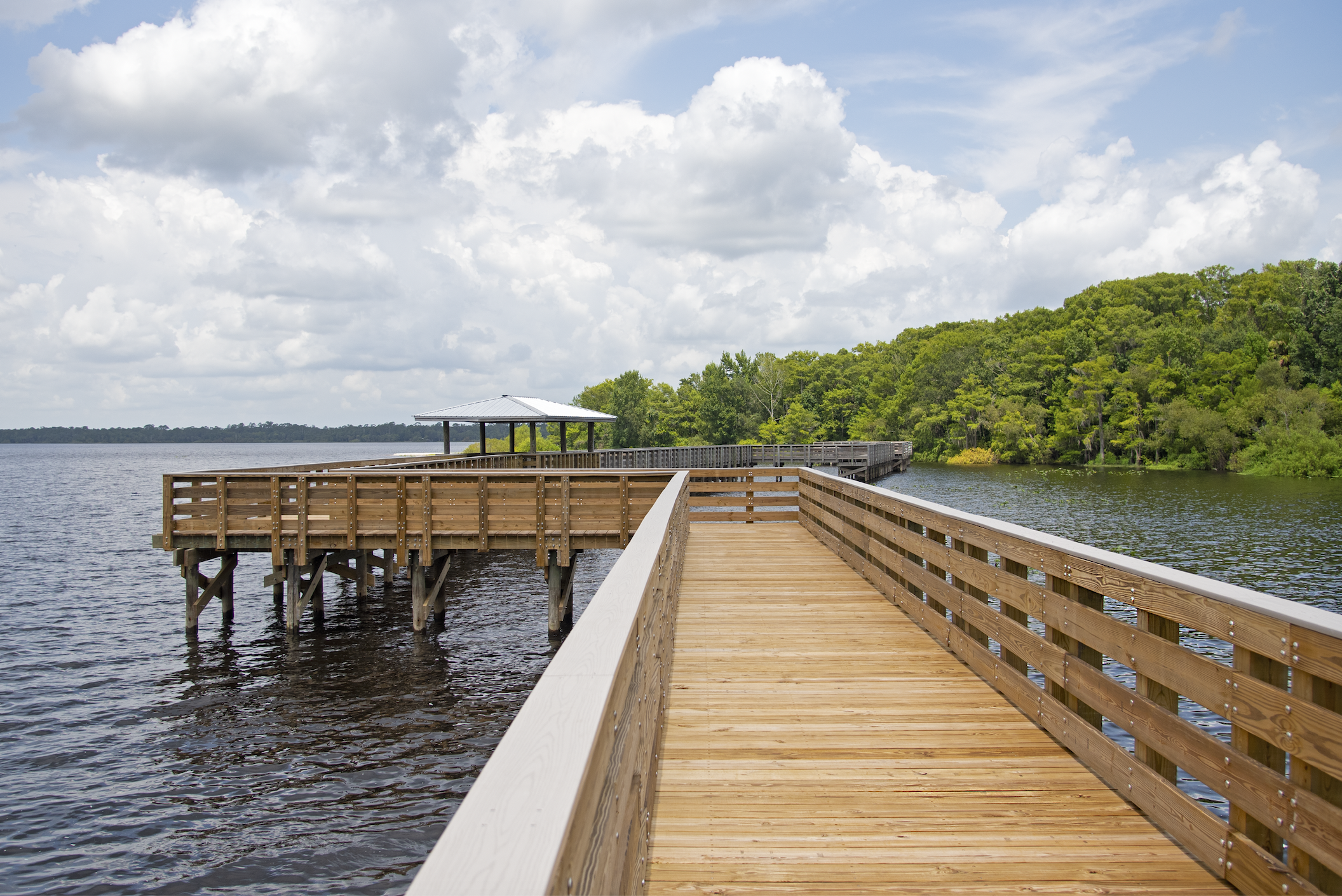 A wooden pier extending over a lake with a small shelter at the end, surrounded by lush green trees and a partly cloudy sky.