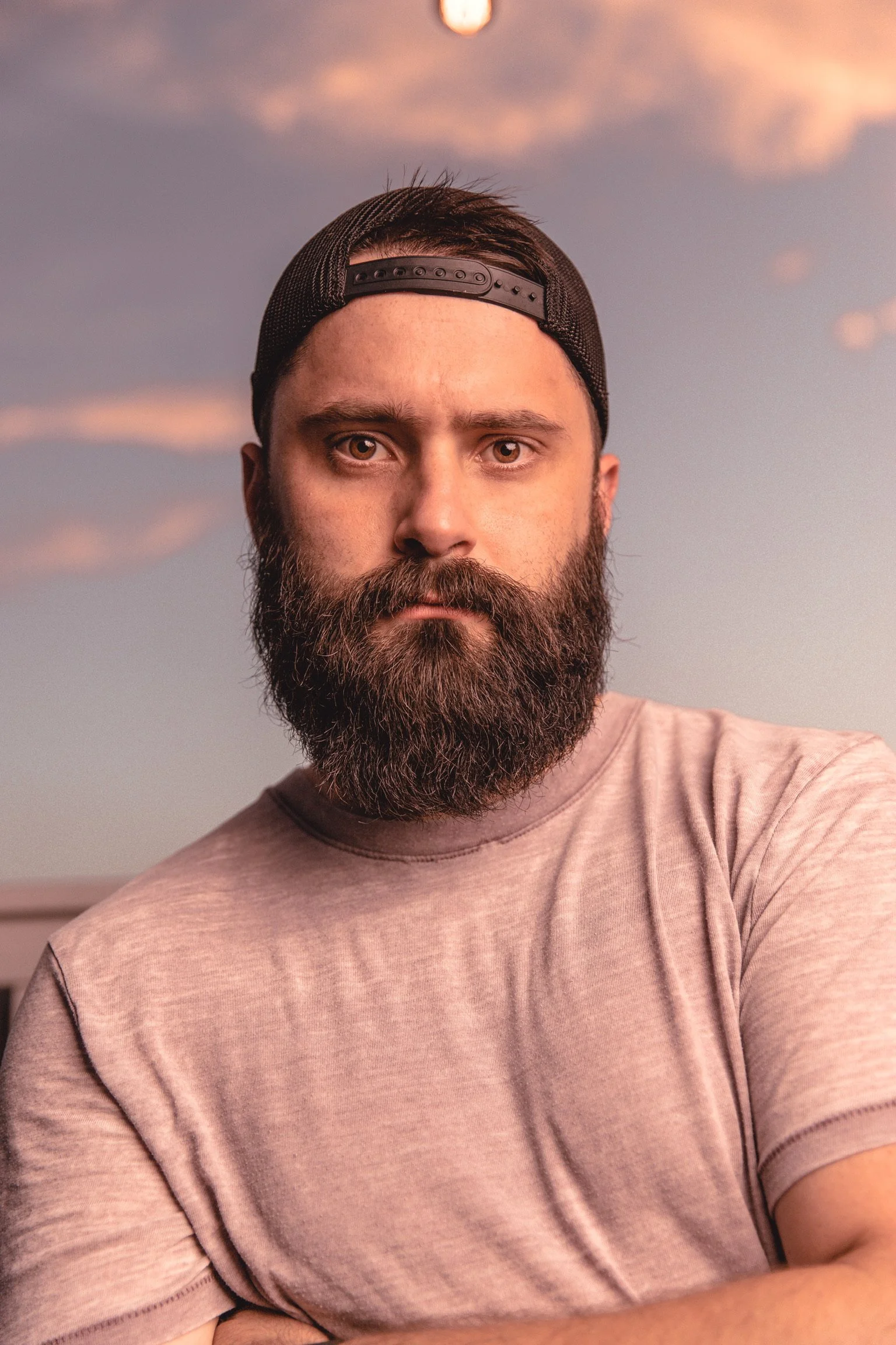 A gay young photographer with a full beard wearing a backwards black cap and a light pink t-shirt, standing outdoors with a sunset sky and scattered clouds in the background.
