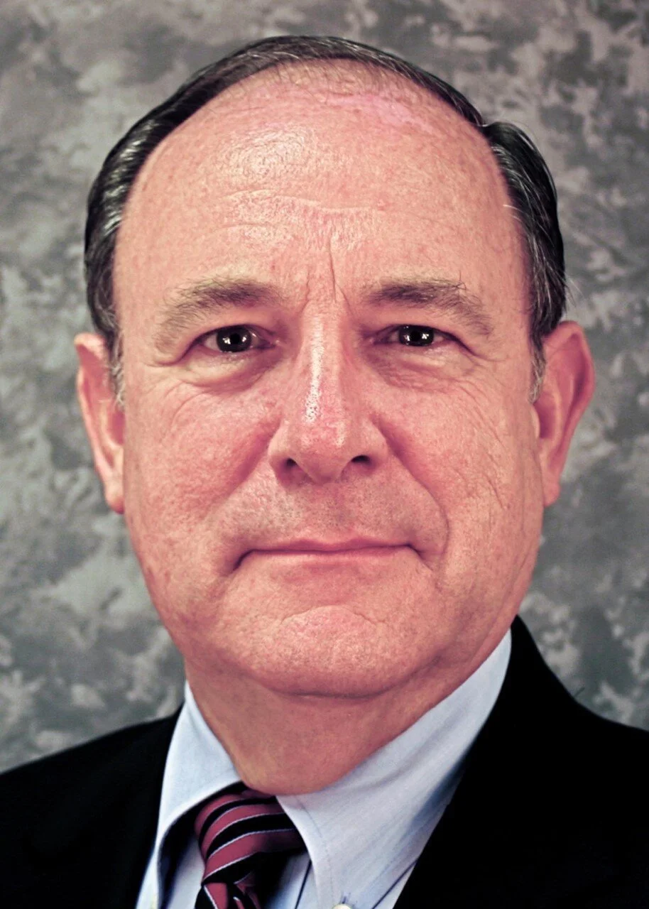 Portrait of an older man with a serious expression, wearing a suit and tie, against a gray textured background.