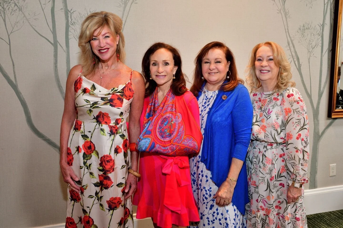 Four women standing together, each wearing brightly colored, floral-patterned dresses, smiling at the camera against a neutral background with subtle tree designs.