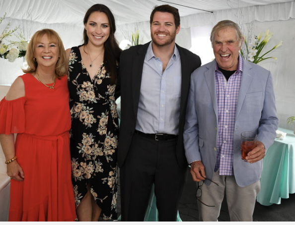 Four smiling people stand together in a well-lit room decorated with white flowers and light-colored furniture. The group includes two women and two men, dressed in formal and semi-formal attire. One woman is wearing a red dress, the other, a floral dress. One man wears a suit, another in a light blazer and checked shirt. A festive atmosphere is noted.