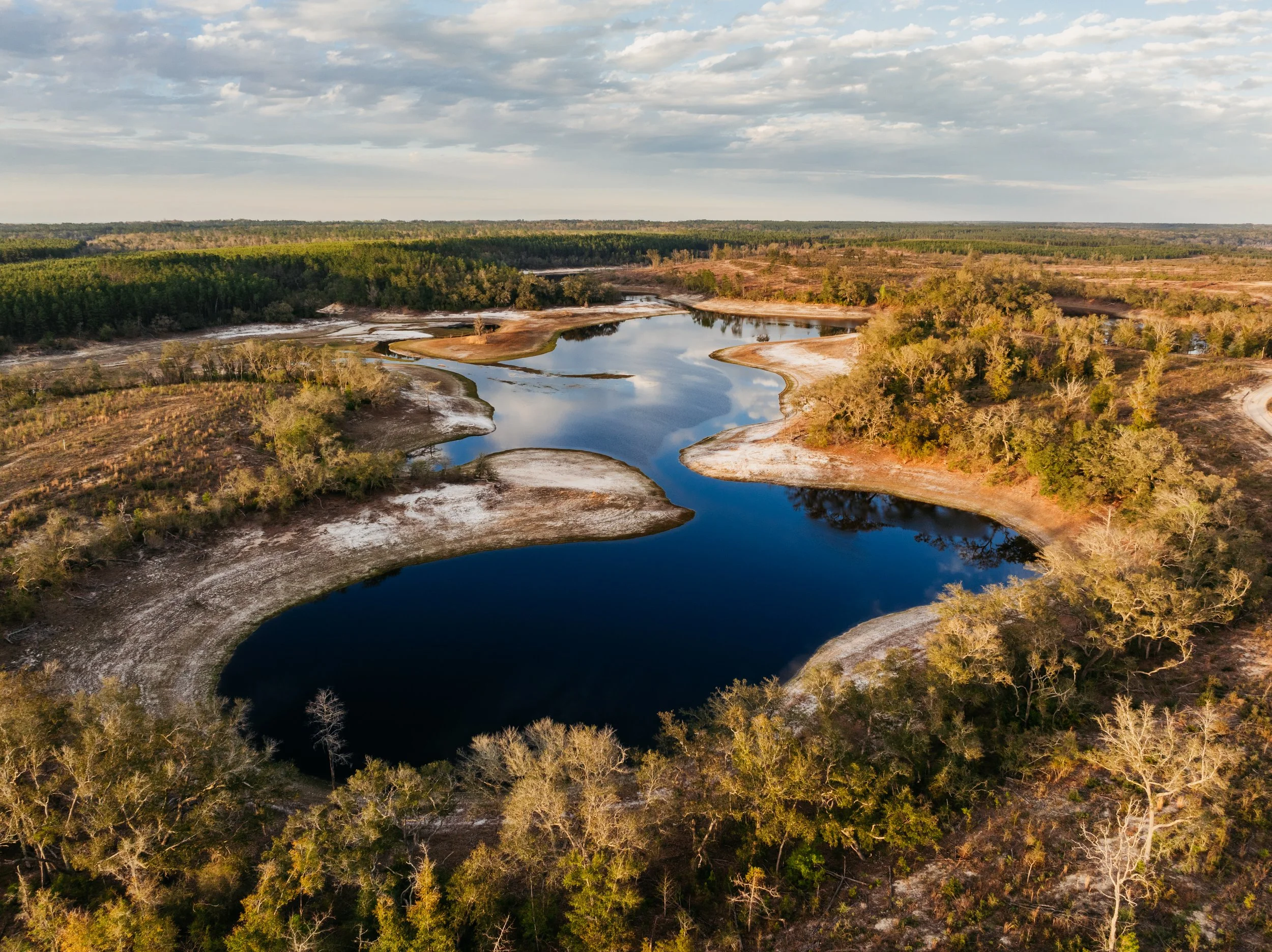 Aerial view of a winding river with dark blue water surrounded by sandy shores and patches of green trees, under a partly cloudy sky.