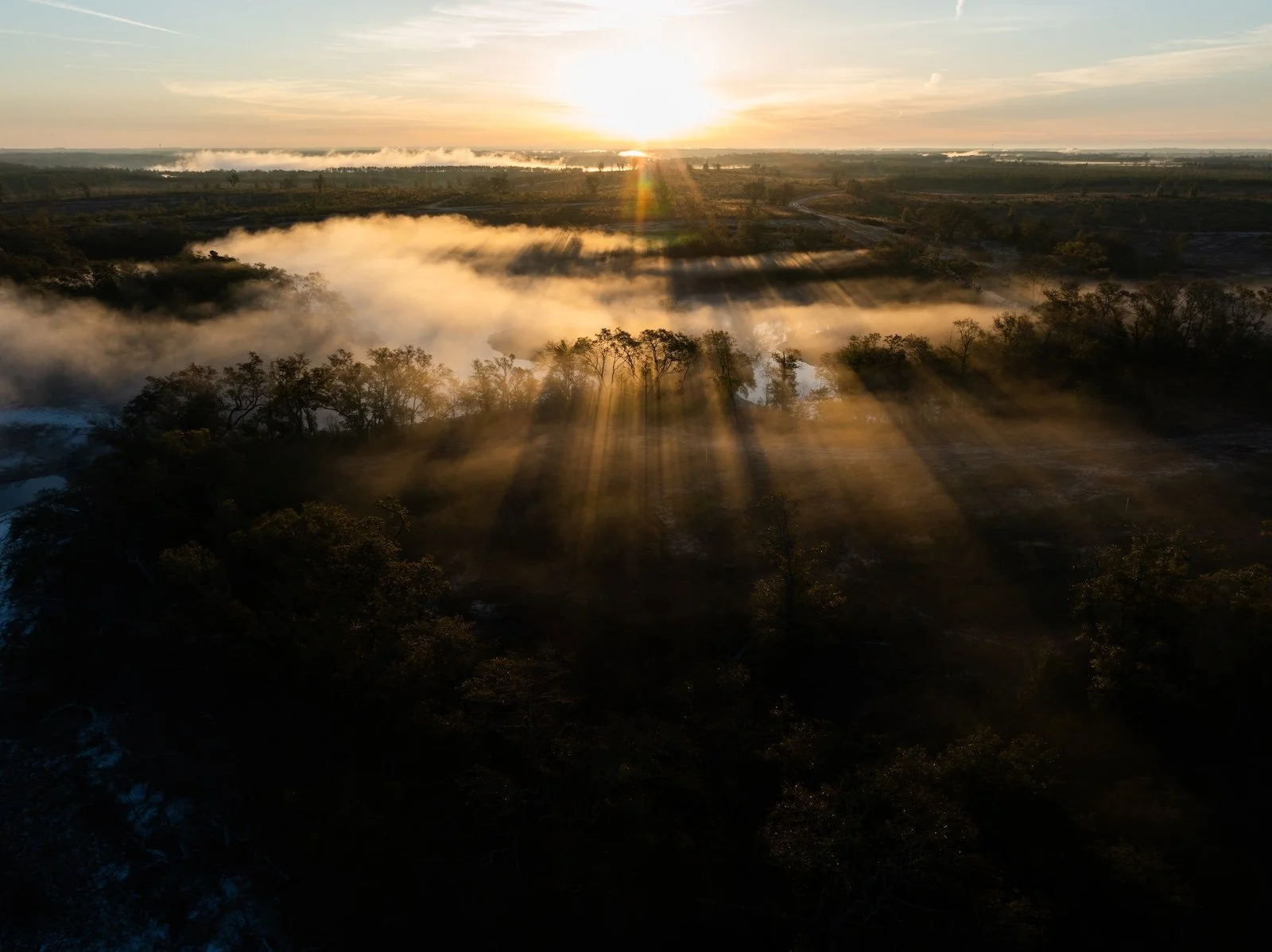 Sun setting over a river with mist and trees, casting long shadows on the landscape.