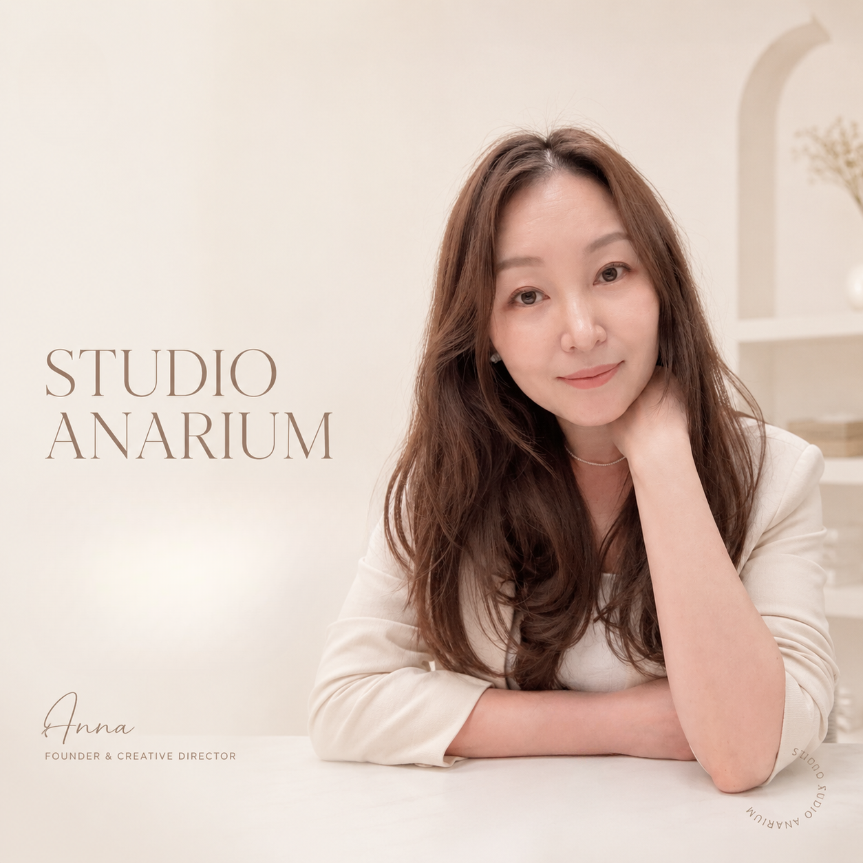 A woman with long brown hair sitting at a white table, resting her chin on her hand, smiling softly. The background features a minimalistic, light-colored interior with shelves and decorative items. Text on the image reads 'STUDIO ANARIUM' and 'Anna, Founder & Creative Director.'