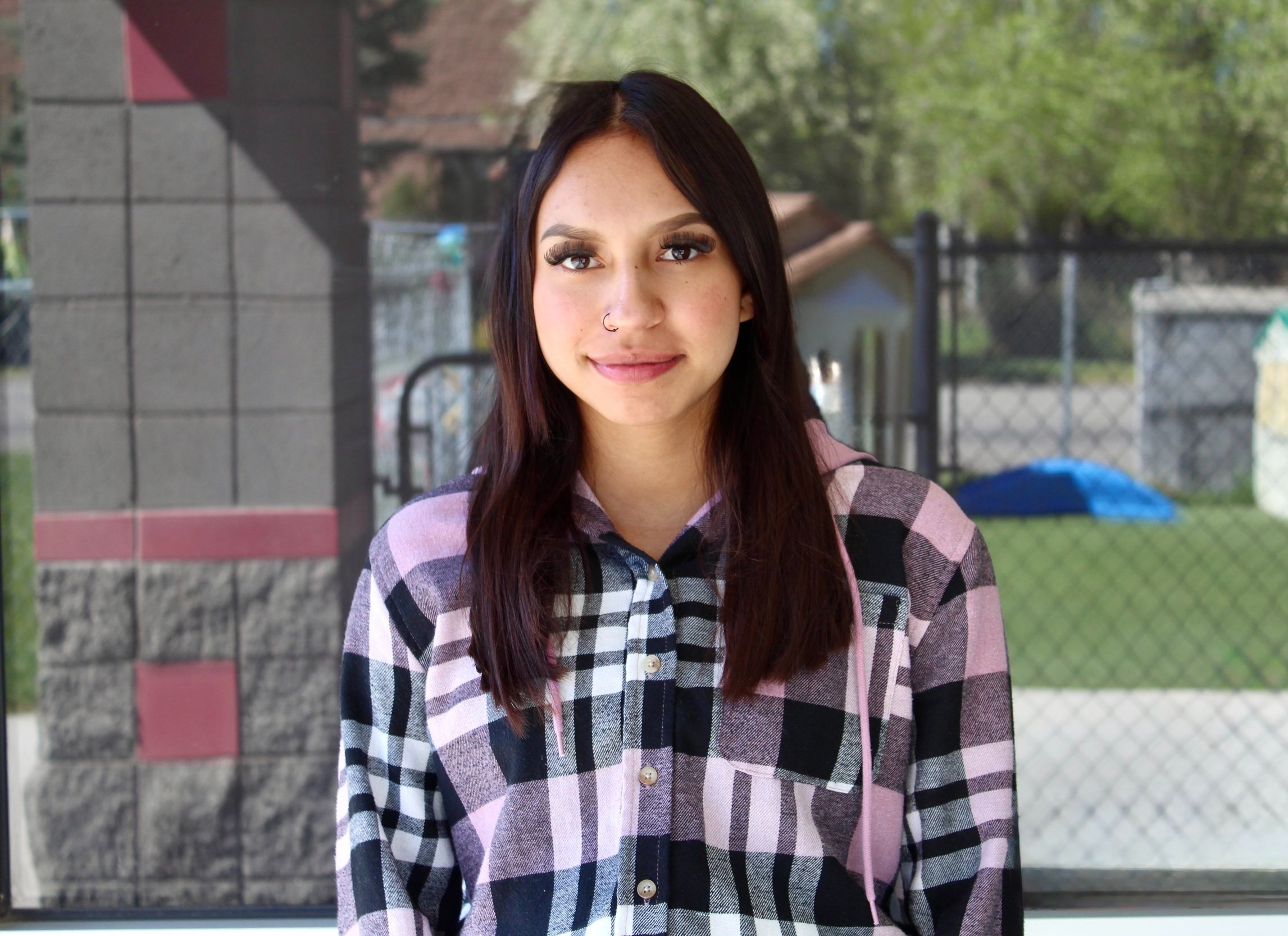 A person with long brown hair wearing a plaid shirt and a nose ring, standing outdoors in front of a fence and greenery.