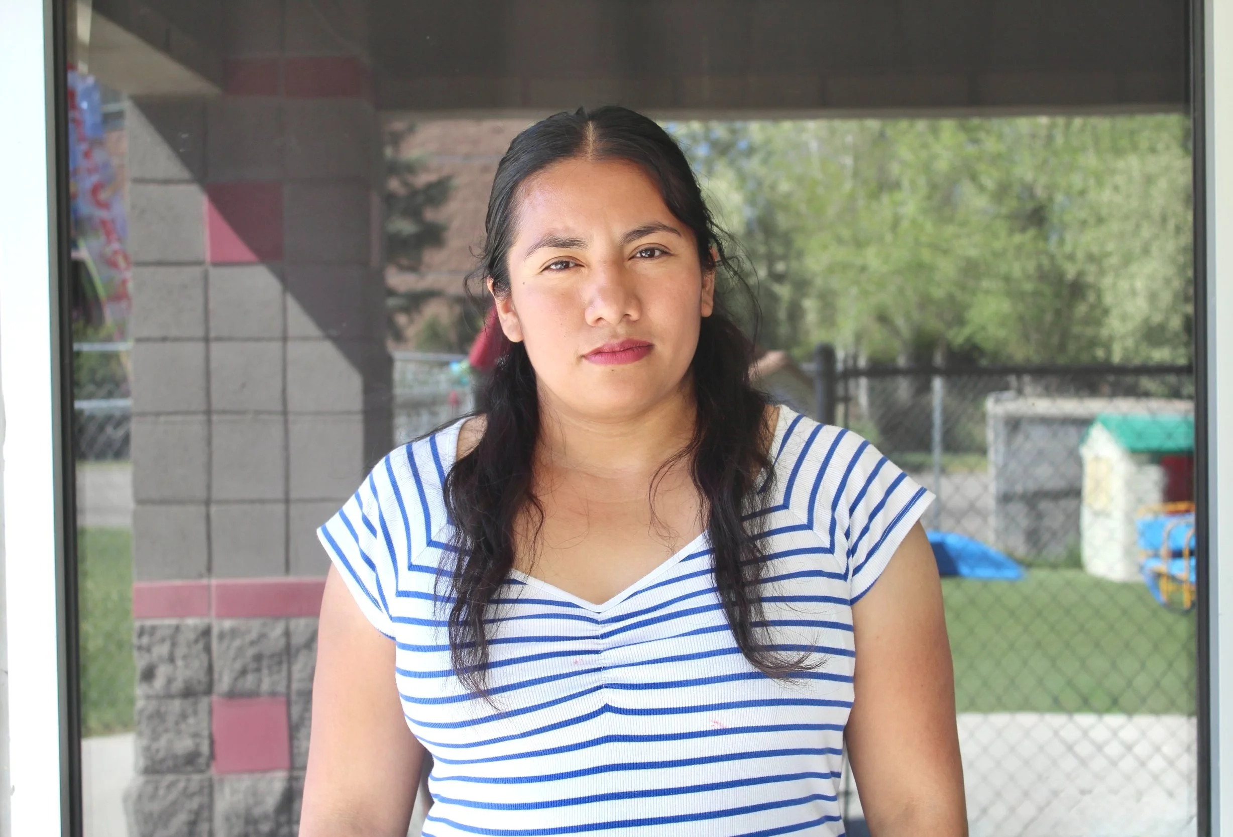 A woman in a striped shirt standing outdoors in front of a fence and playground.