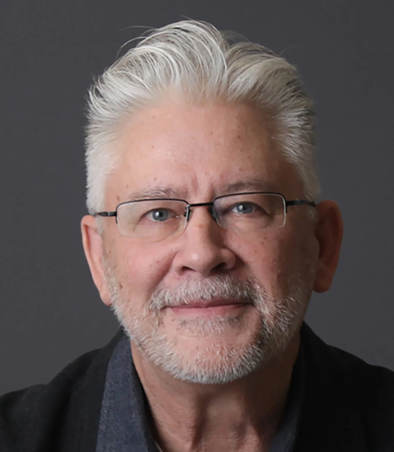 Close-up portrait of an older man with white hair, glasses, and a beard, wearing a dark blazer and a collared shirt against a plain dark background.