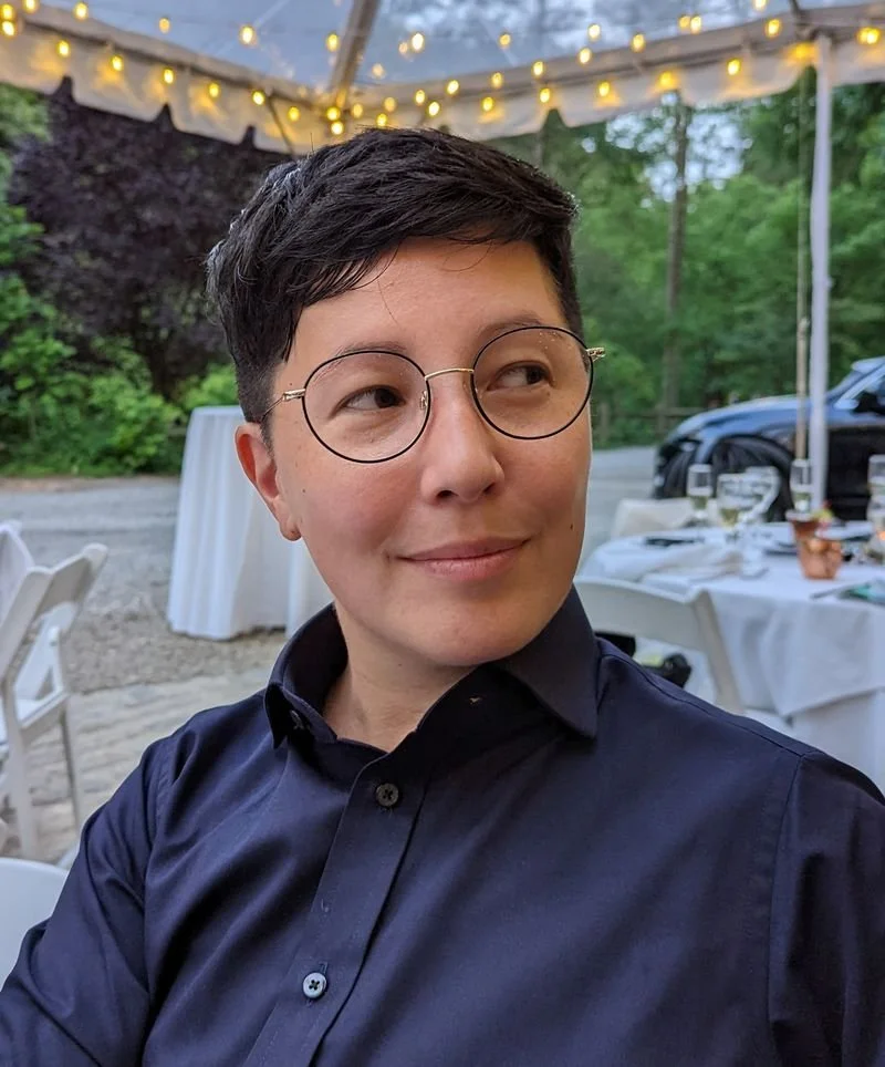 A person with short dark hair and glasses smiling at an outdoor event under a tent with string lights, surrounded by trees and decorated tables.