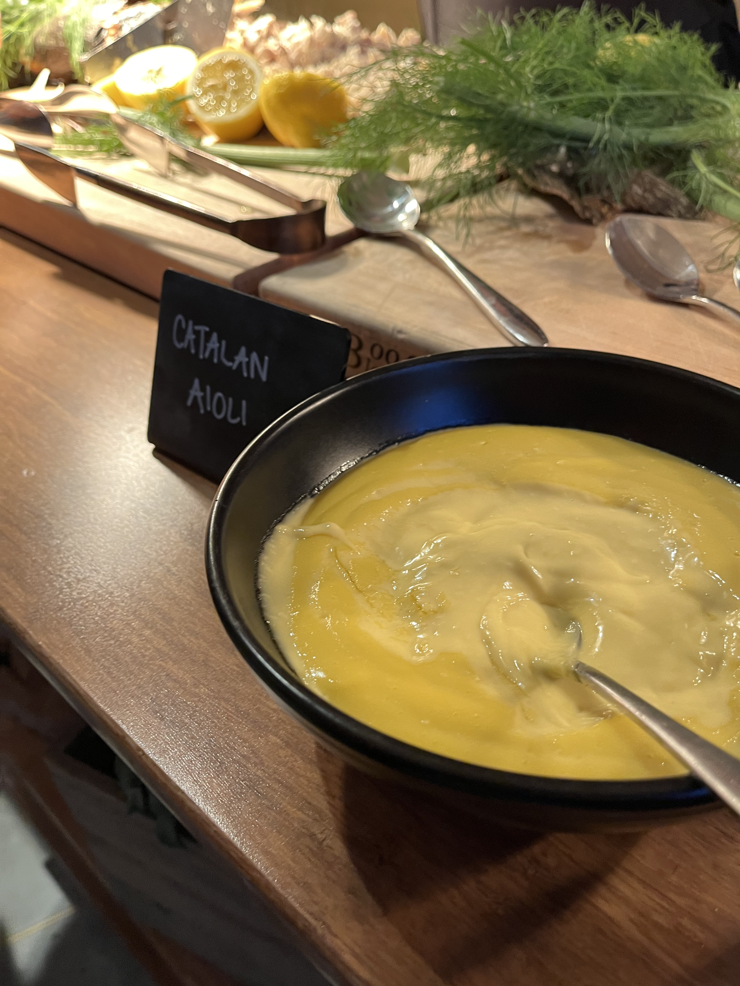 A black bowl of creamy, yellow, cilantro aloi with a spoon in it on a wooden table. In the background, there are lemon halves, herbs, and serving utensils, with a small black sign reading 'Cilantro Aioli'.