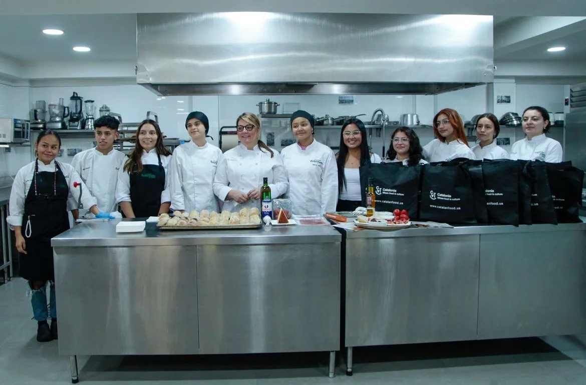 Group of culinary students and chefs standing behind a stainless steel kitchen counter with baked goods and ingredients, in a commercial kitchen.