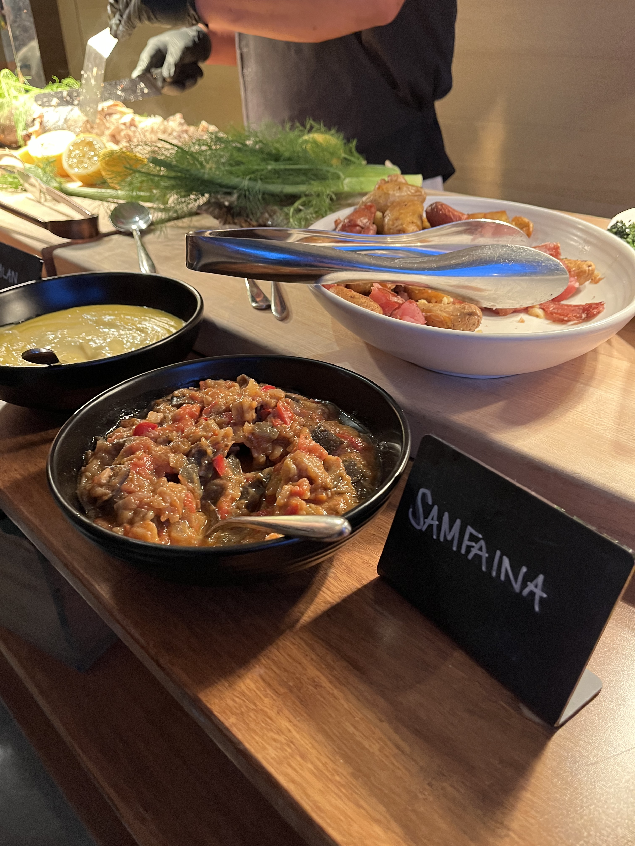 A buffet with a bowl of eggplant dip, a bowl of yellow sauce, and a large white bowl of assorted breakfast meats including sausages, bacon, and ham. A person is seen in the background serving themselves.