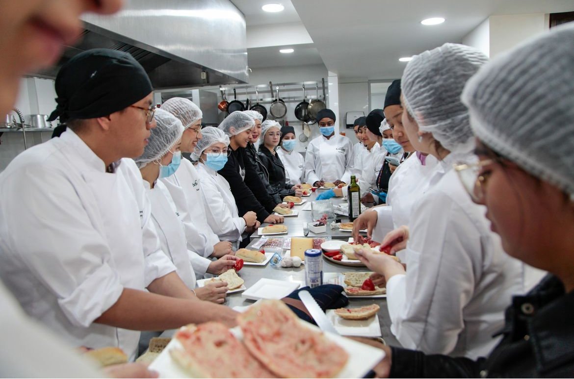 Group of chefs and culinary students wearing uniforms and hairnets around a countertop, preparing sandwiches in a commercial kitchen.