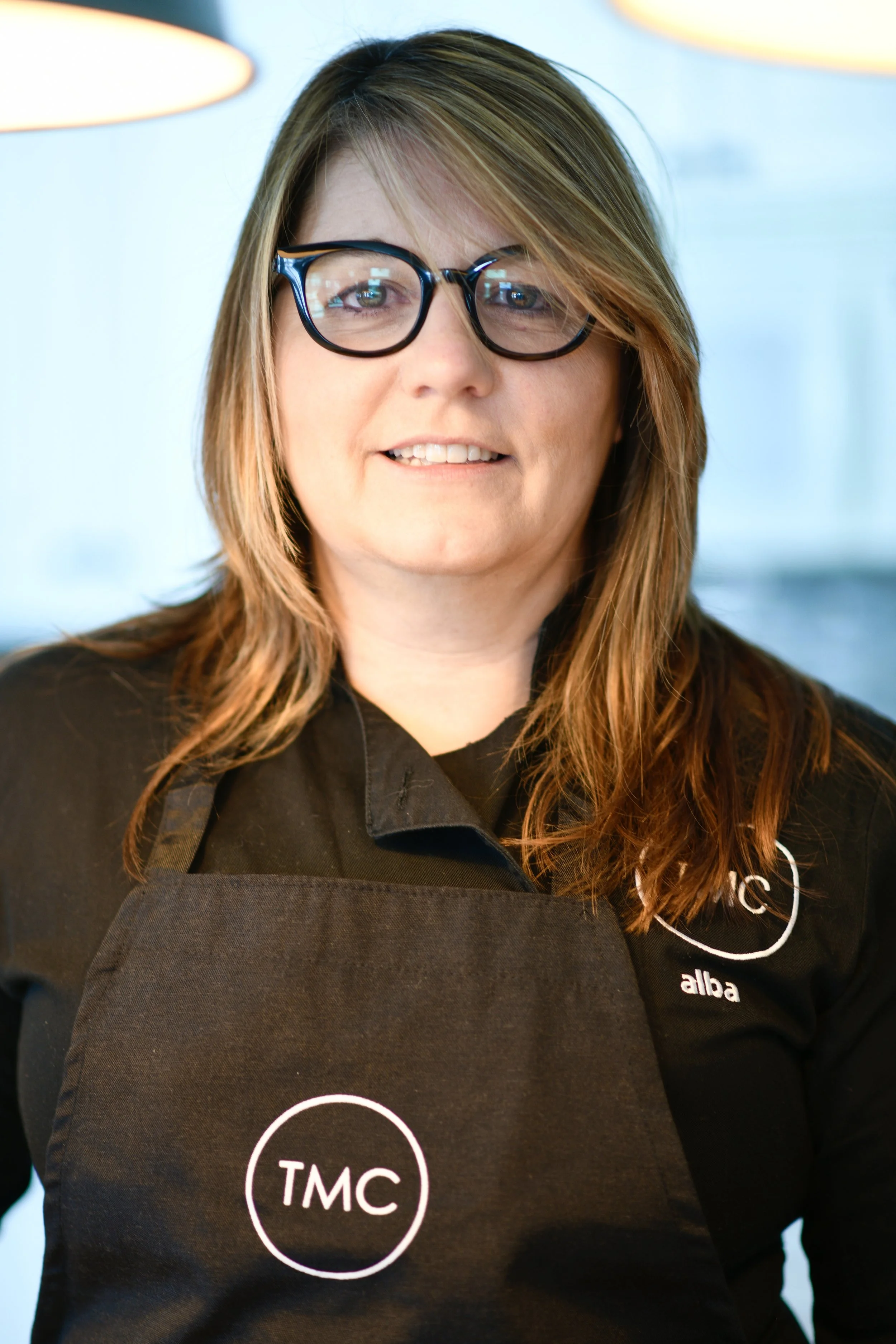 A woman with glasses and shoulder-length hair wearing a black apron with the TMC logo, smiling indoors.
