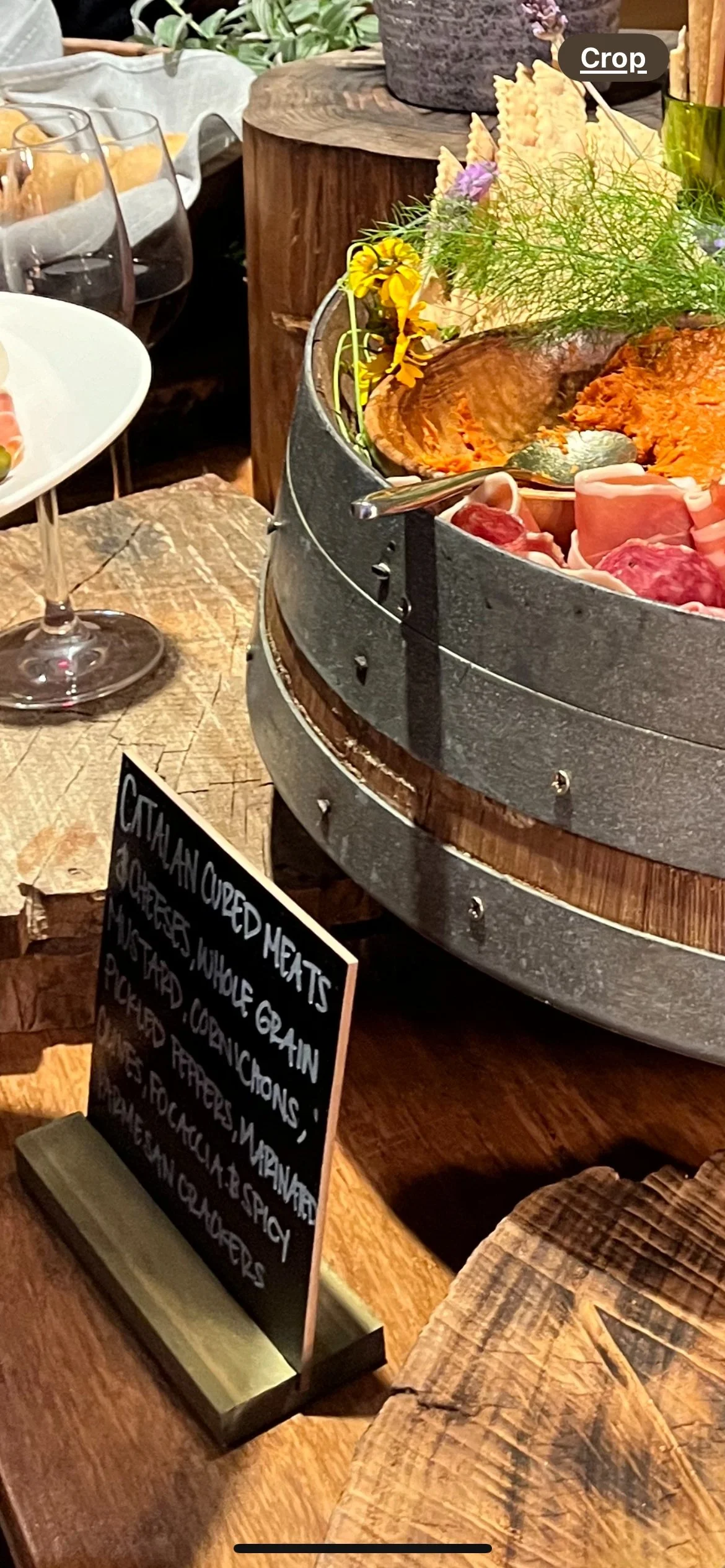 A rustic wooden table displays a large metal and wood steamer basket filled with assorted cooked meats, garnished with fresh herbs and flowers. A small chalkboard sign indicates the dish is smoked meats, including pork and beef.