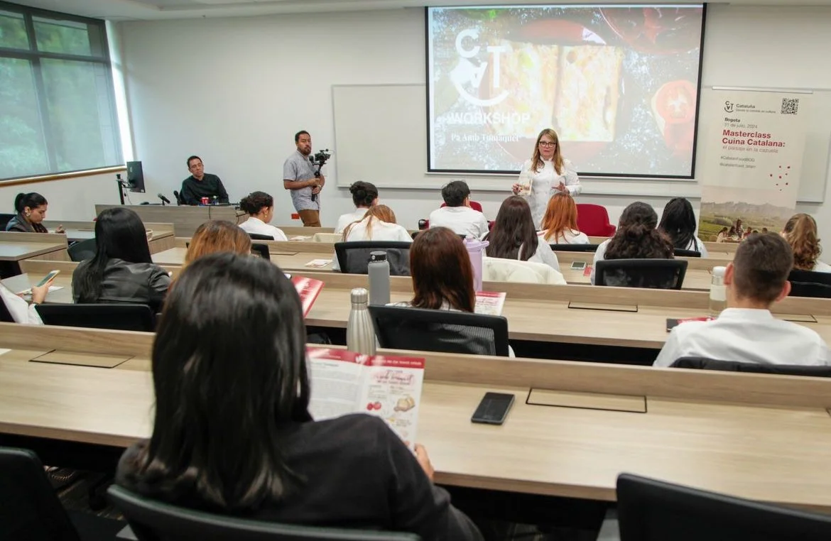 A woman is giving a presentation in a classroom or conference room, with a large screen behind her displaying a colorful image and text. Attendees are seated at tables, some taking notes or looking at materials, while one person is filming the session with a camera.