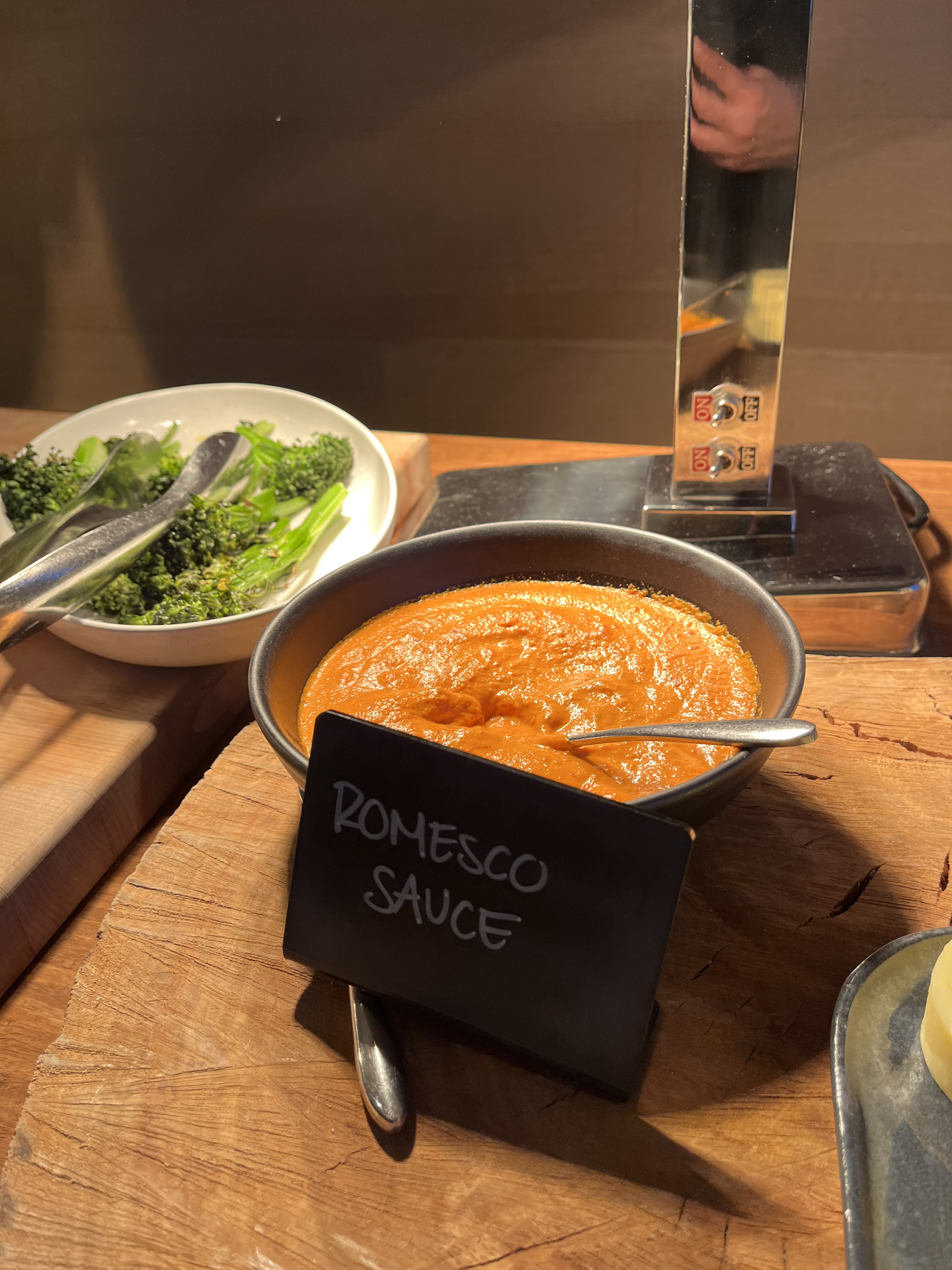 A small black bowl of orange romesco sauce with a spoon inside, placed on a round wooden cutting board. A black chalkboard sign in front of the bowl reads 'ROMESCO SAUCE'. To the left, there is a white bowl filled with broccoli and other greens, and tongs are resting inside it. There is a metal grill or heat source behind the sauce.