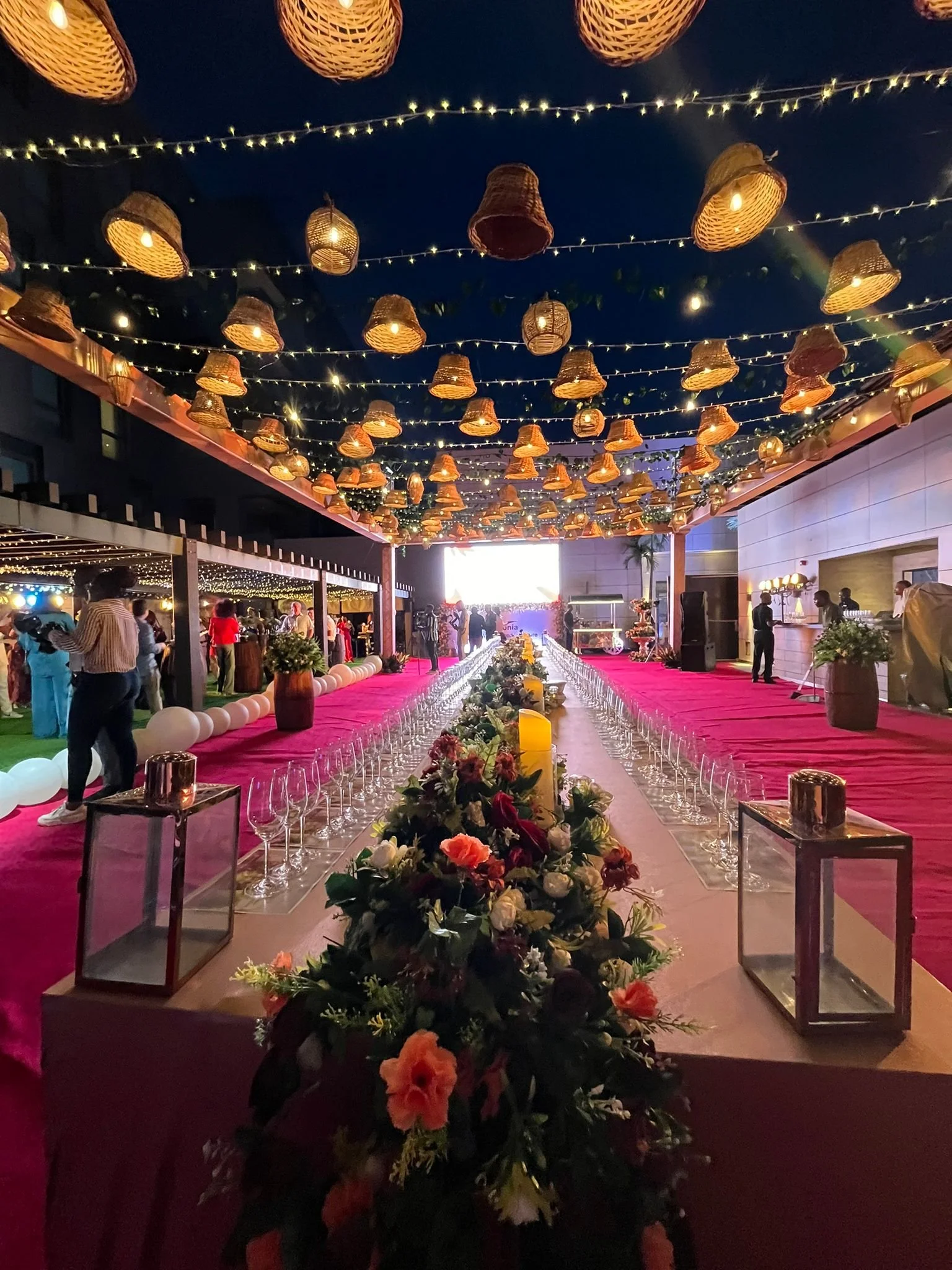 A decorated event space with hanging wicker lanterns and fairy lights, a long table with flowers and candles, and people mingling in the background.
