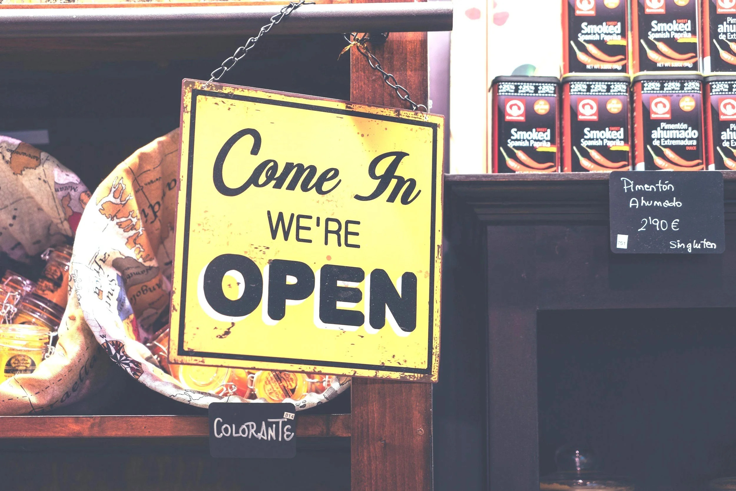 A yellow sign hanging on a chain saying 'Come In We're OPEN' next to shelves with Spanish smoked paprika and other products.
