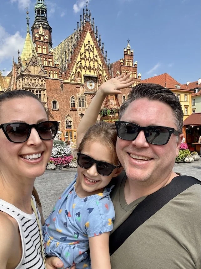 A smiling family of three wearing sunglasses takes a selfie in front of a historic building with gothic architecture, decorative towers, and a clock, during the daytime.