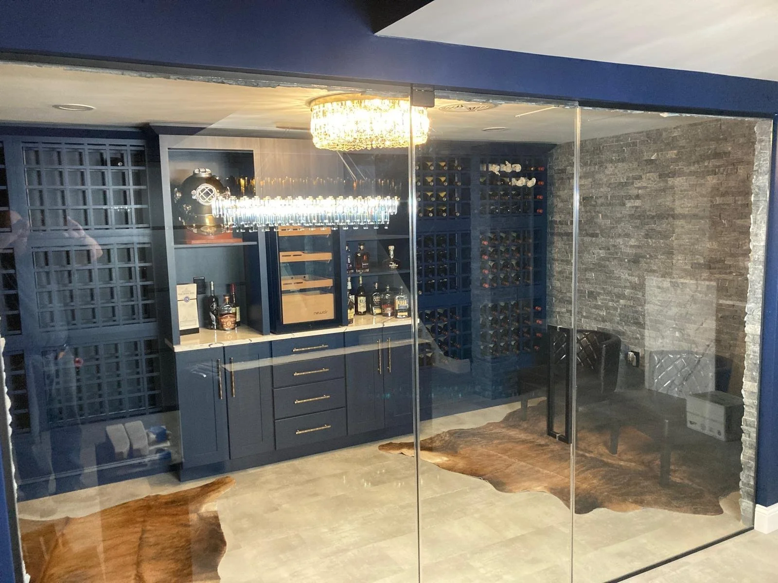 View through glass window of a home bar with blue cabinets, wine bottles stored in racks, a wine cooler, liquor bottles, a chandelier, and a seating area with a rug and a black chair.