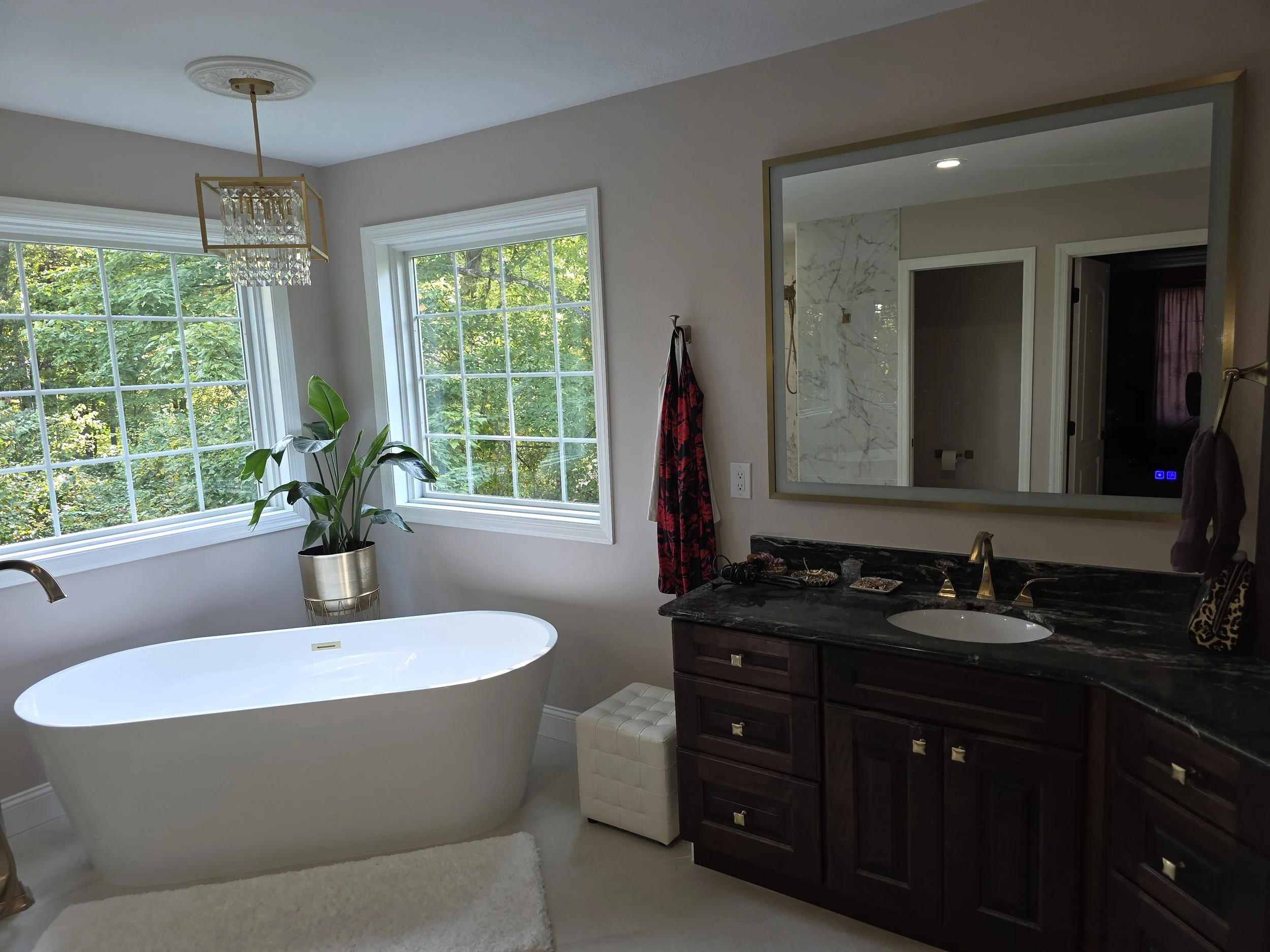 A bathroom with a freestanding bathtub near a window, a plant in a gold pot, a large mirror, a dark wood vanity with a granite countertop, a small white padded stool, a towel hanging on a hook, and a marble shower wall visible in the reflection.