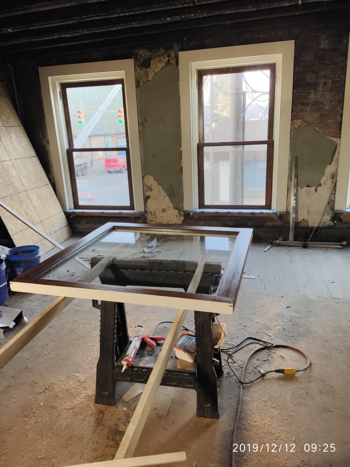 Interior of a room under renovation with two windows, some construction tools, and a worktable with a glass piece. The walls are partially stripped, showing exposed brick and plaster.