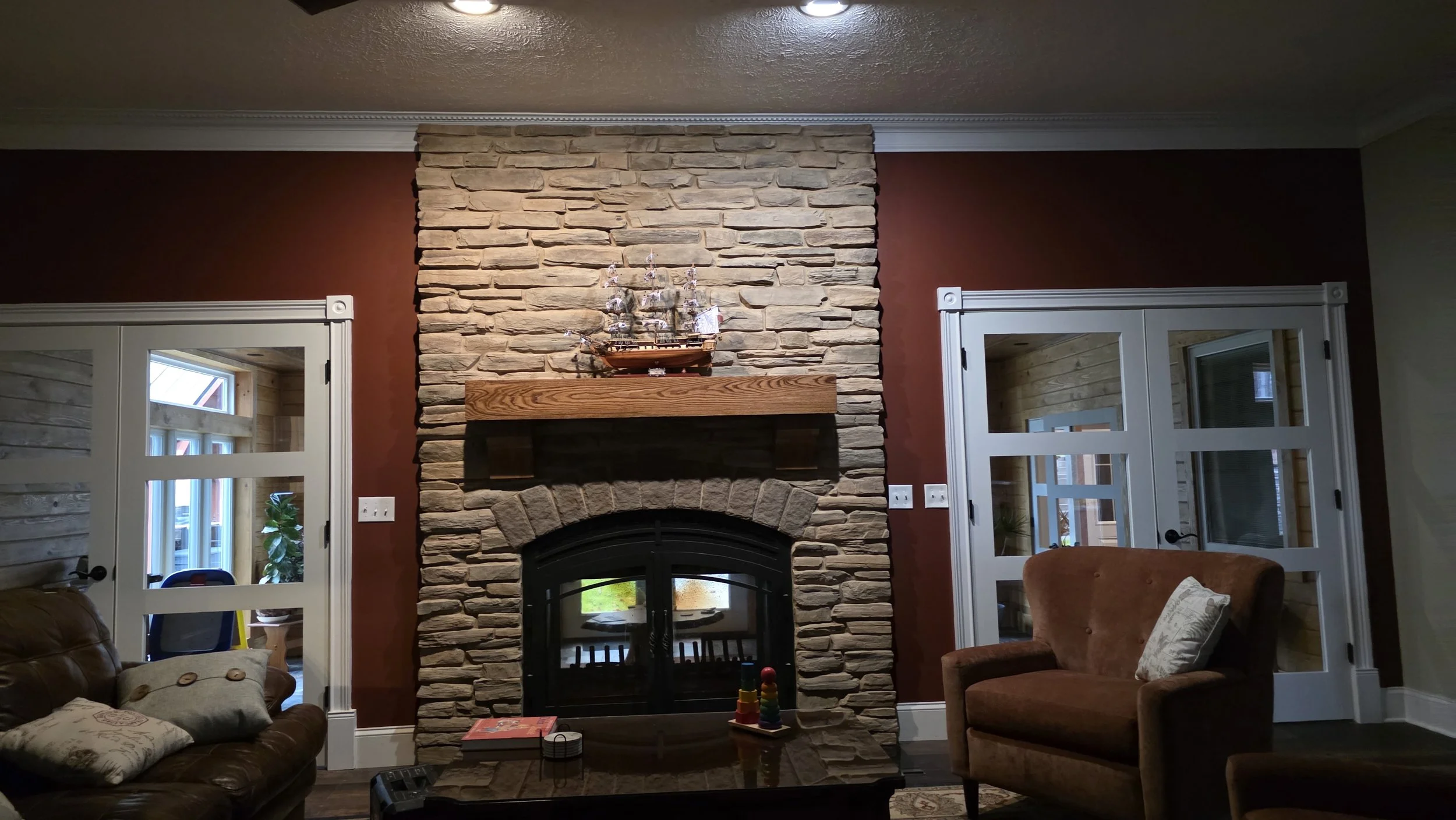 Living room with a stone fireplace, wooden mantel, and two white French doors. There is a brown leather couch with pillows, an orange armchair, and a coffee table.