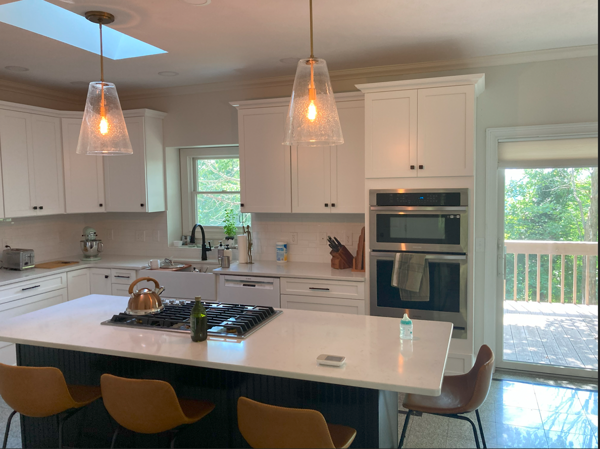 Modern kitchen with white cabinets, a central island with a stove and a teapot, pendant lights, a window with greenery outside, and a sliding glass door leading to a deck.