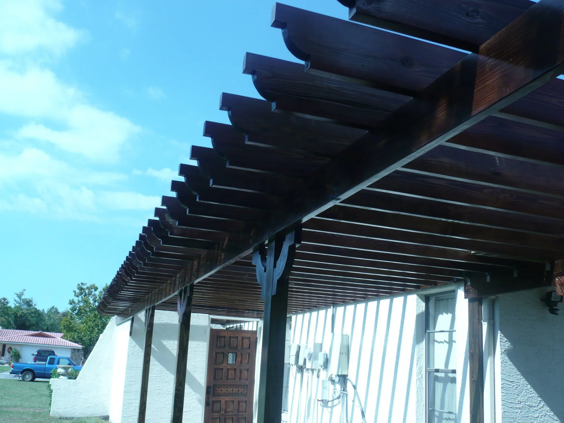 Wooden pergola attached to a white building with a brown door, under a blue sky with some clouds, in a residential area.