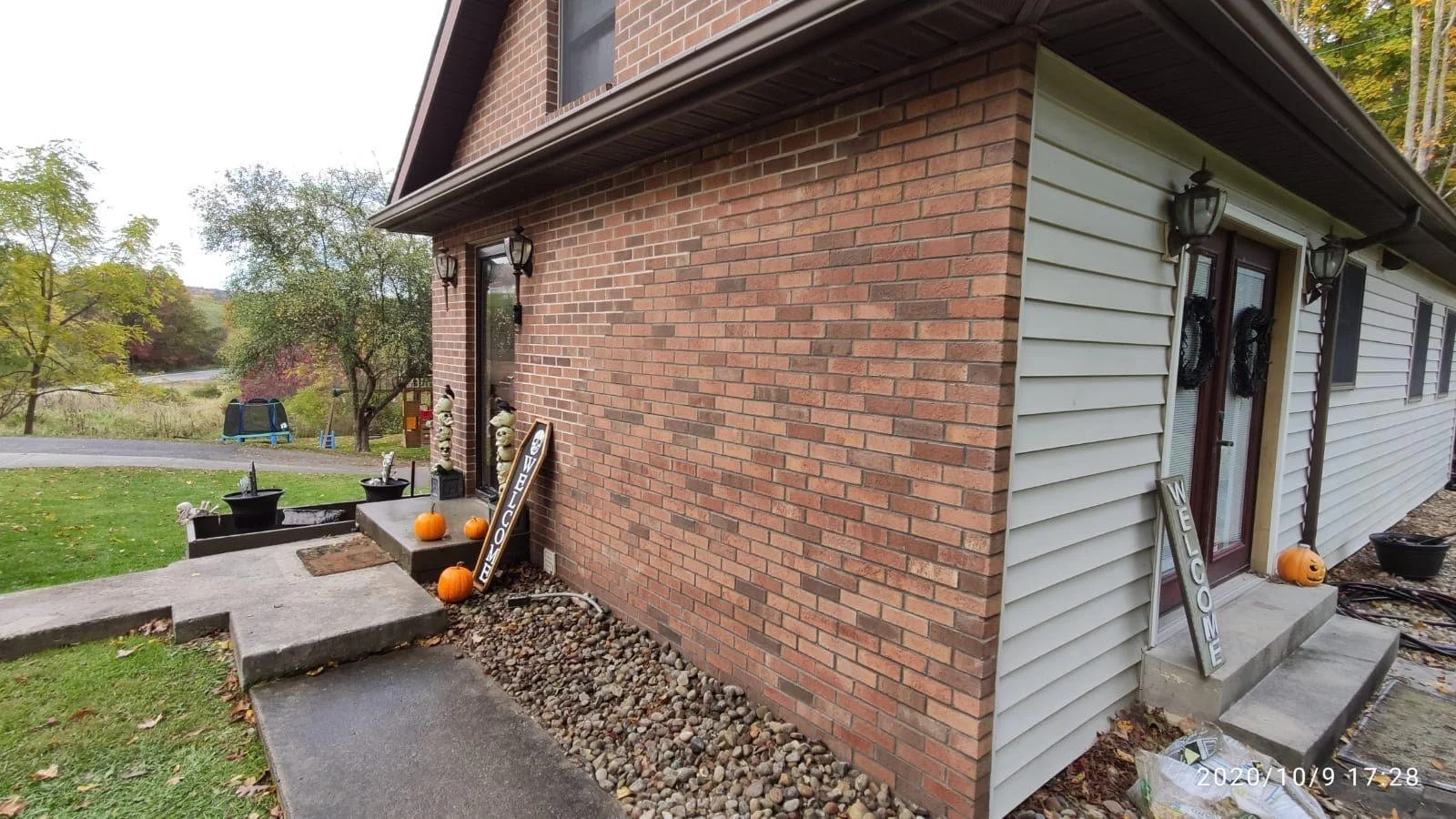 Front porch of a house decorated for fall with pumpkins, welcome signs, and wreaths on the door, surrounded by trees and a lawn.