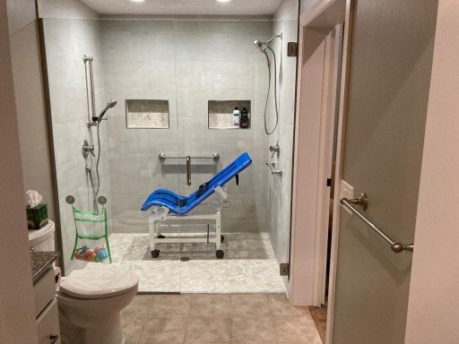 Accessible bathroom with a walk-in shower and a blue plastic chair. The shower has grab bars, two built-in wall niches, and handheld showerheads. Toilet and towel bars are visible, with a green plastic chair beside the toilet.