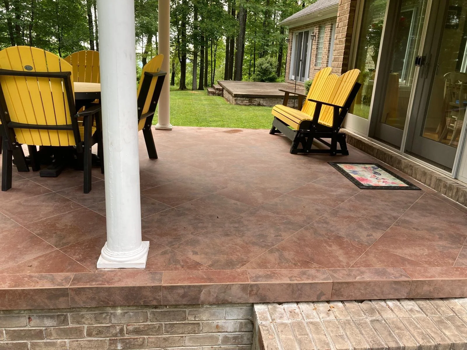 A patio with brown tile flooring, yellow wooden chairs, a yellow wooden bench near a sliding glass door, and a white pillar, with a grassy backyard and wooded area in the background.