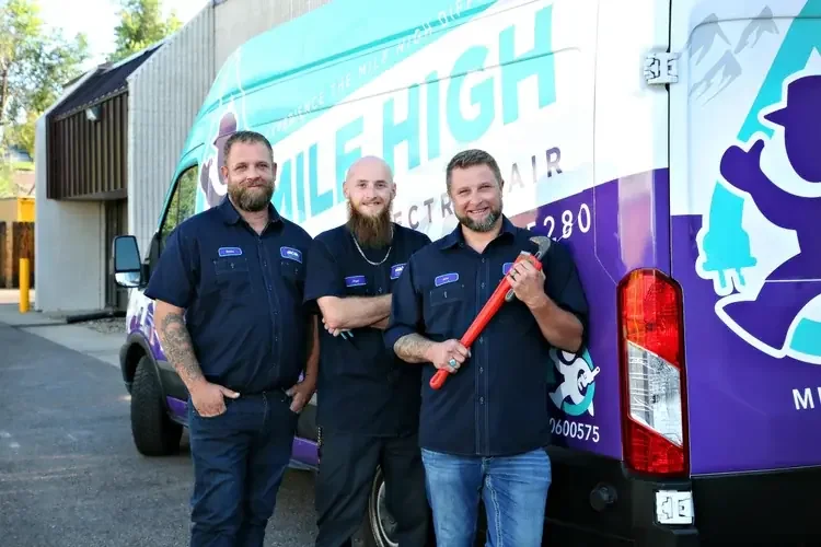 Three men standing in front of an electric repair vehicle branded with 'Mile High Electric' and a logo. They are wearing matching dark blue uniforms, with one holding a large red pipe wrench. The background includes a building and trees.