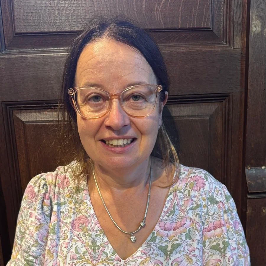 A woman with dark hair, glasses, and a floral top smiling in front of a wooden door.
