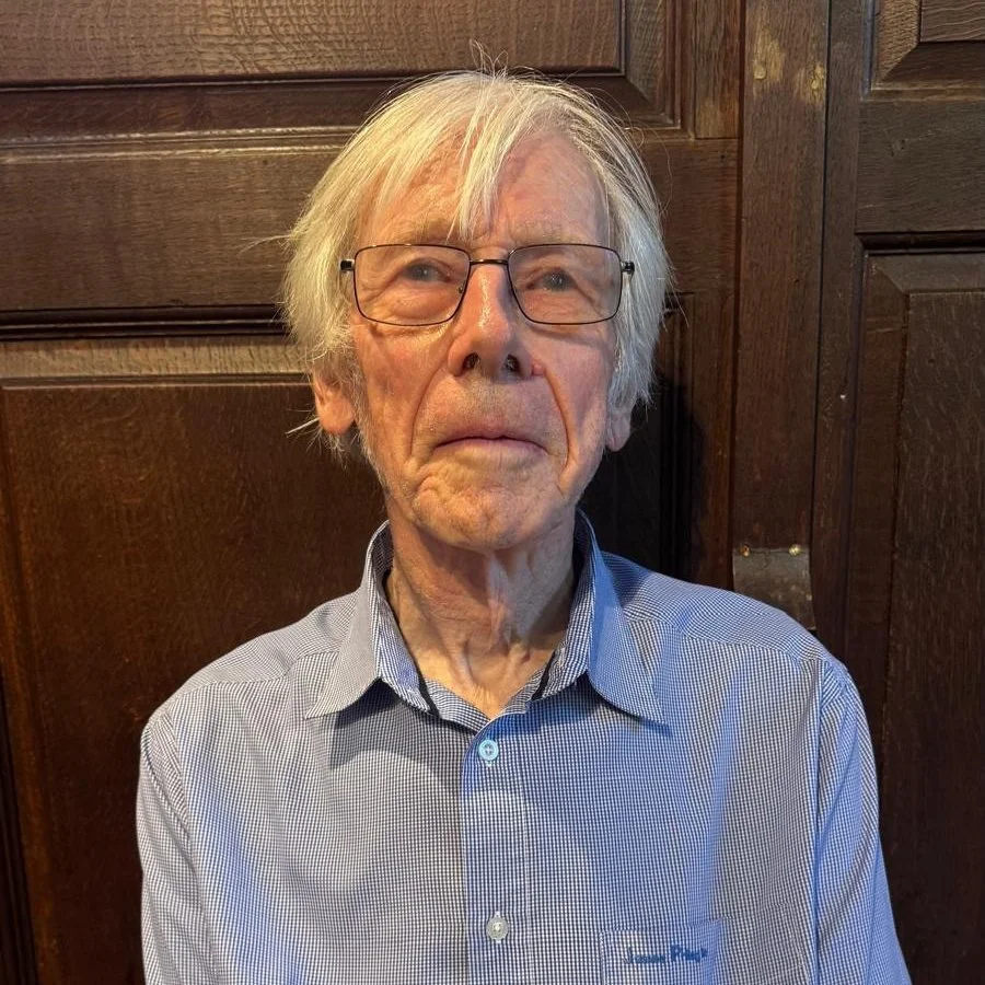 An elderly man with white hair, glasses, and a checkered shirt standing in front of a wooden wall.