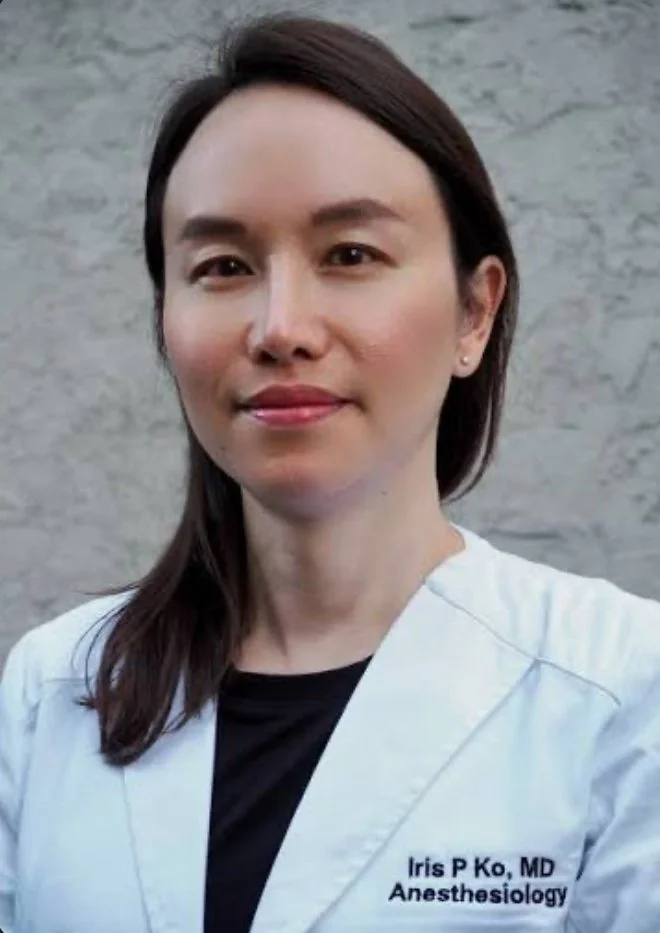 A woman in a white medical coat standing outdoors against a gray stone wall, looking at the camera and smiling gently.