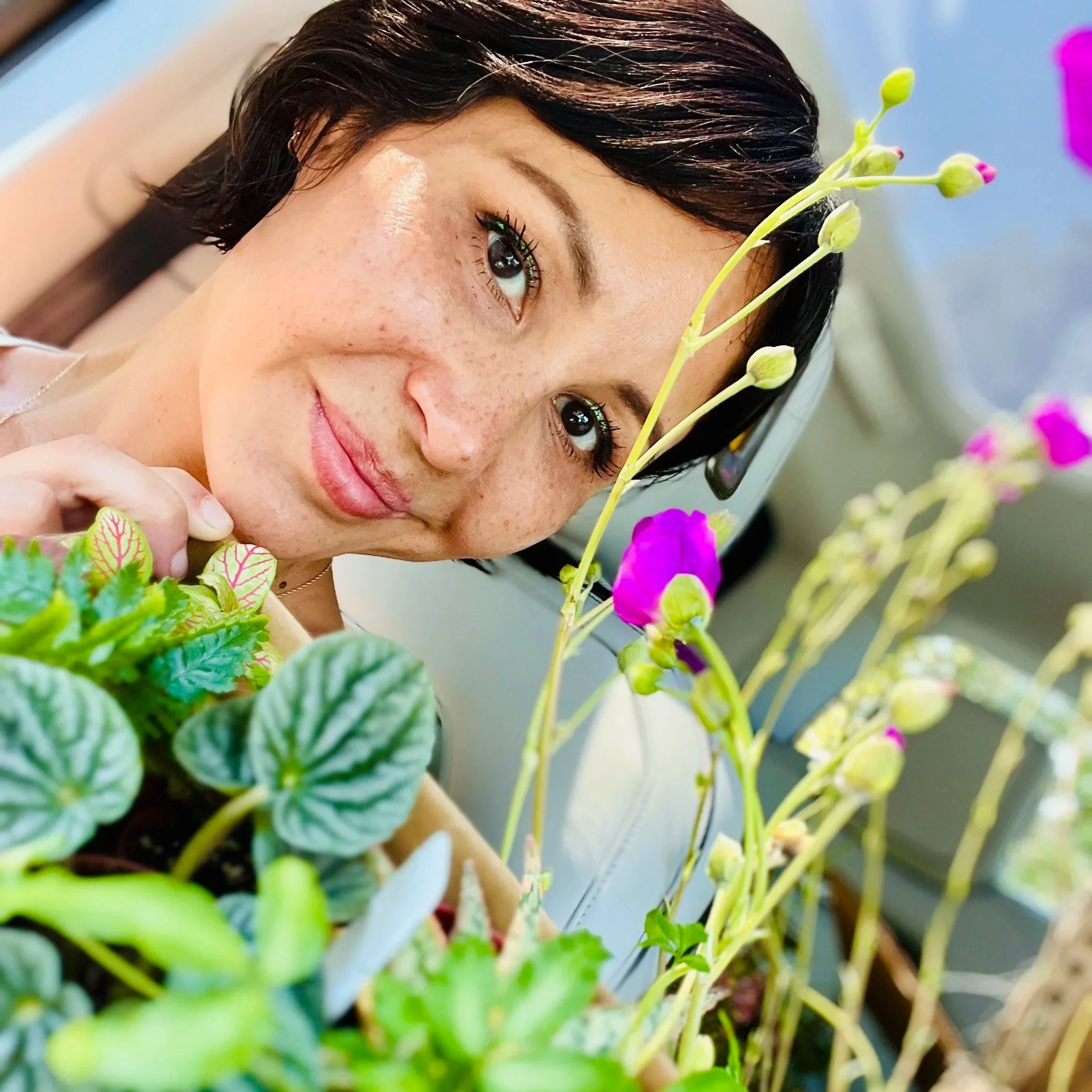 A woman with short dark hair, freckles, and long eyelashes leaning over potted plants with colorful flowers and green foliage, indoors near a window.