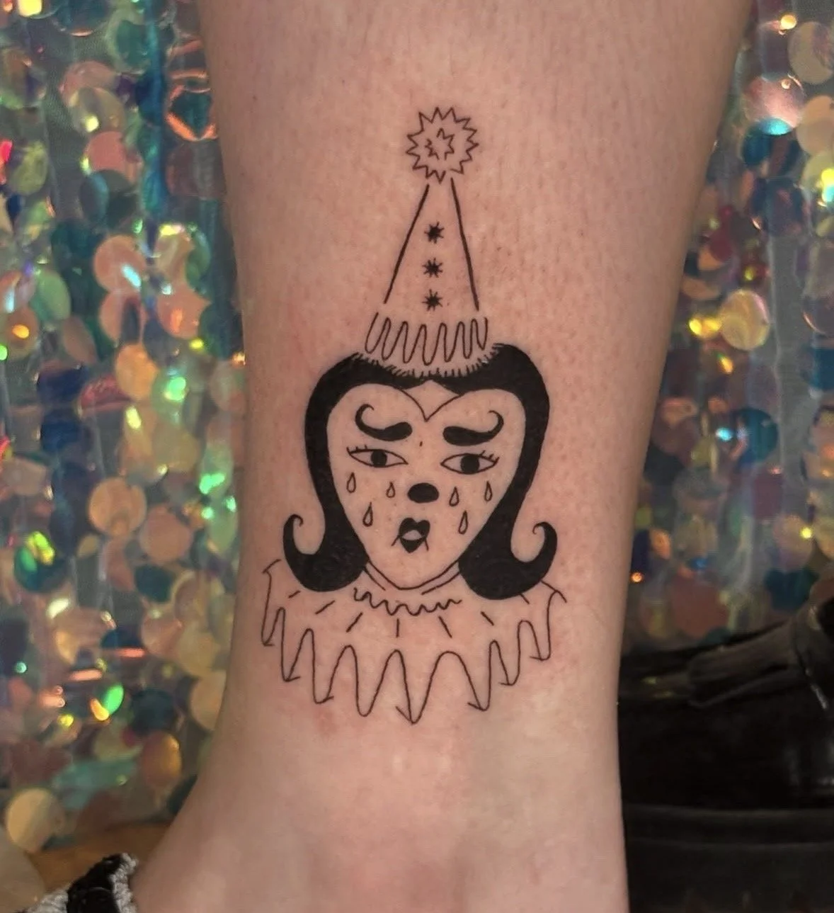 A tattoo of a crying clown with a sad expression, tears, and a clown costume with a ruffled collar. The clown has black hair and eyebrows, and a small nose. Above the clown's head is a simple, stylized hat with stars and a burst at the top, giving a 