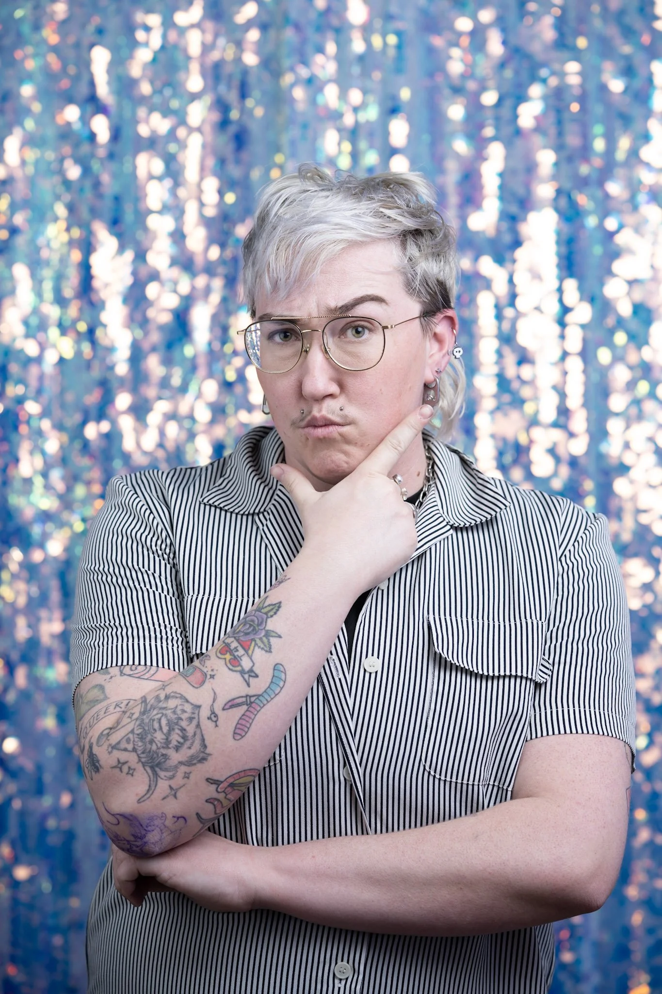 Person with platinum blonde hair, glasses, and tattoos on left arm, wearing a striped shirt, posing with one hand on chin against a colorful, sparkly background.