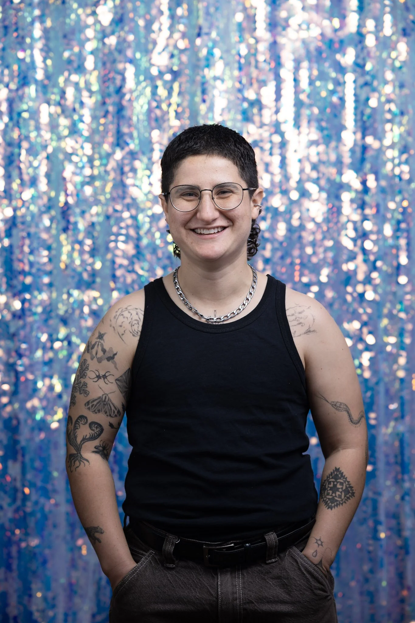 A person with short dark hair and tattoos on their arms, wearing glasses, a silver chain necklace, a black sleeveless shirt, and dark pants, stands in front of a glittery, colorful backdrop, smiling.