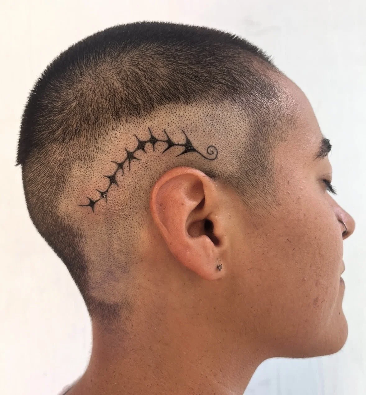 Side profile of a person with short, shaved hair and a decorative tattoo behind the ear.