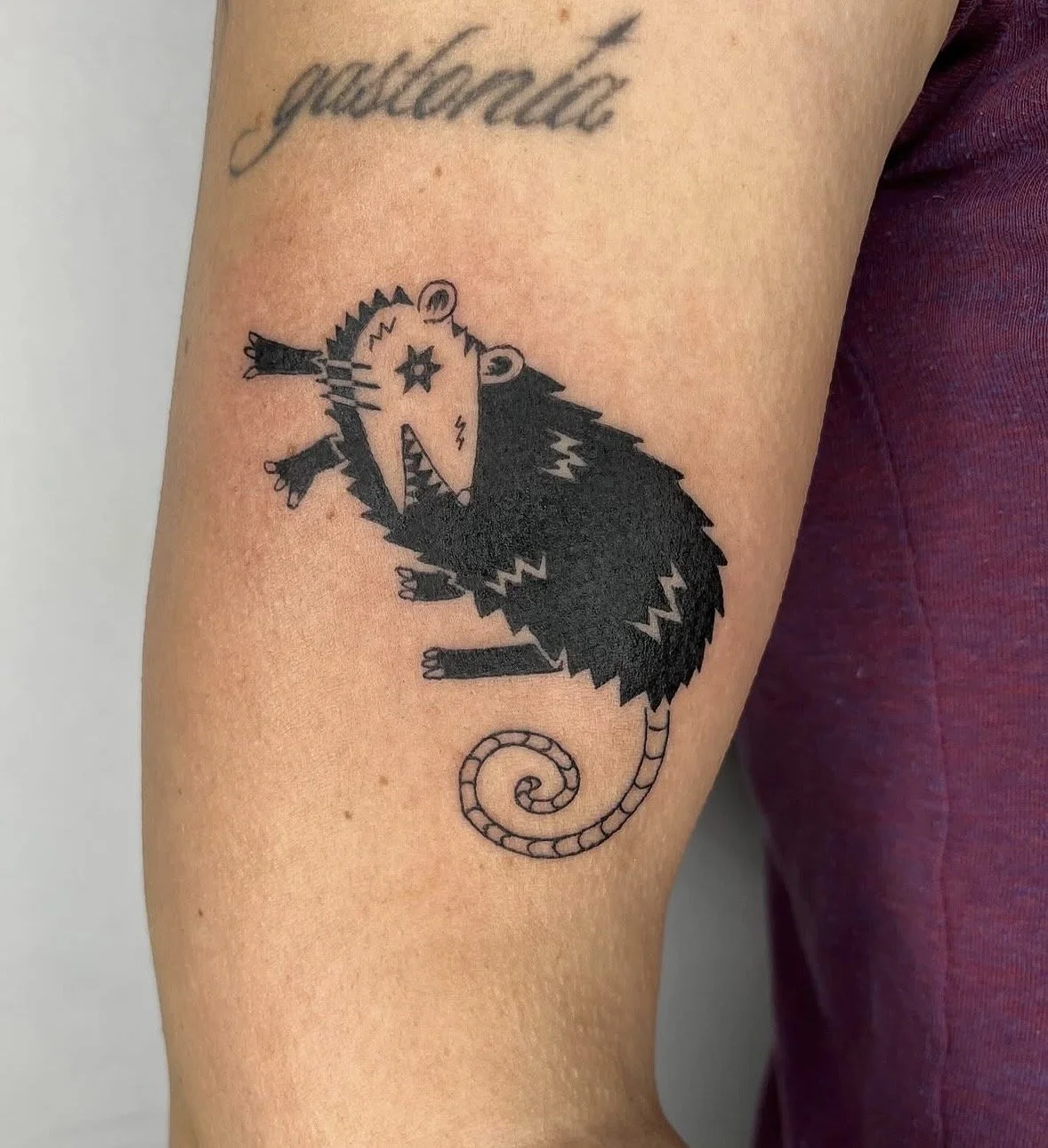 Tattoo of a stylized rat with a star on its eye, sharp teeth, and a curled tail on arm.