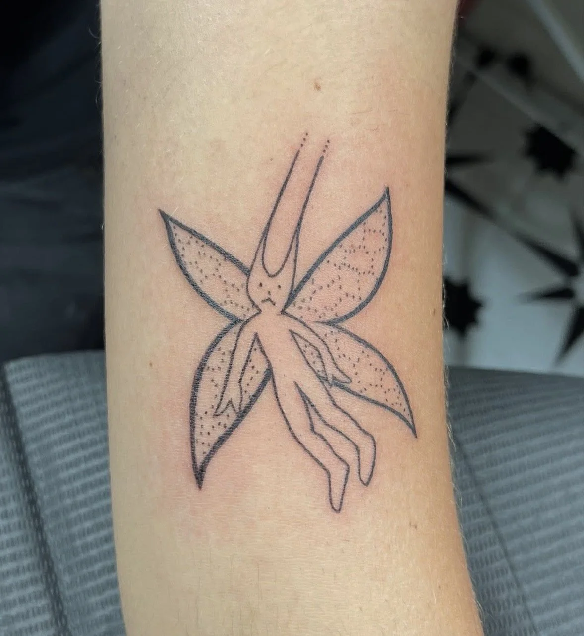 A tattoo of a stylized fairy with wings and long hair, featuring dot shading and outline details.