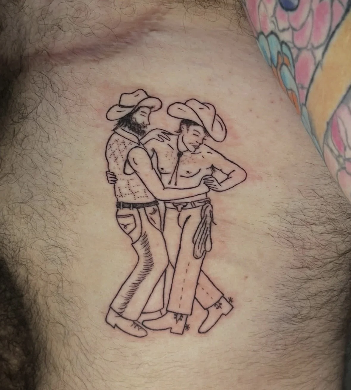 Tattoo of two men in cowboy hats and jeans, hugging and smiling, with one shirtless and the other wearing a fitted shirt and trousers.