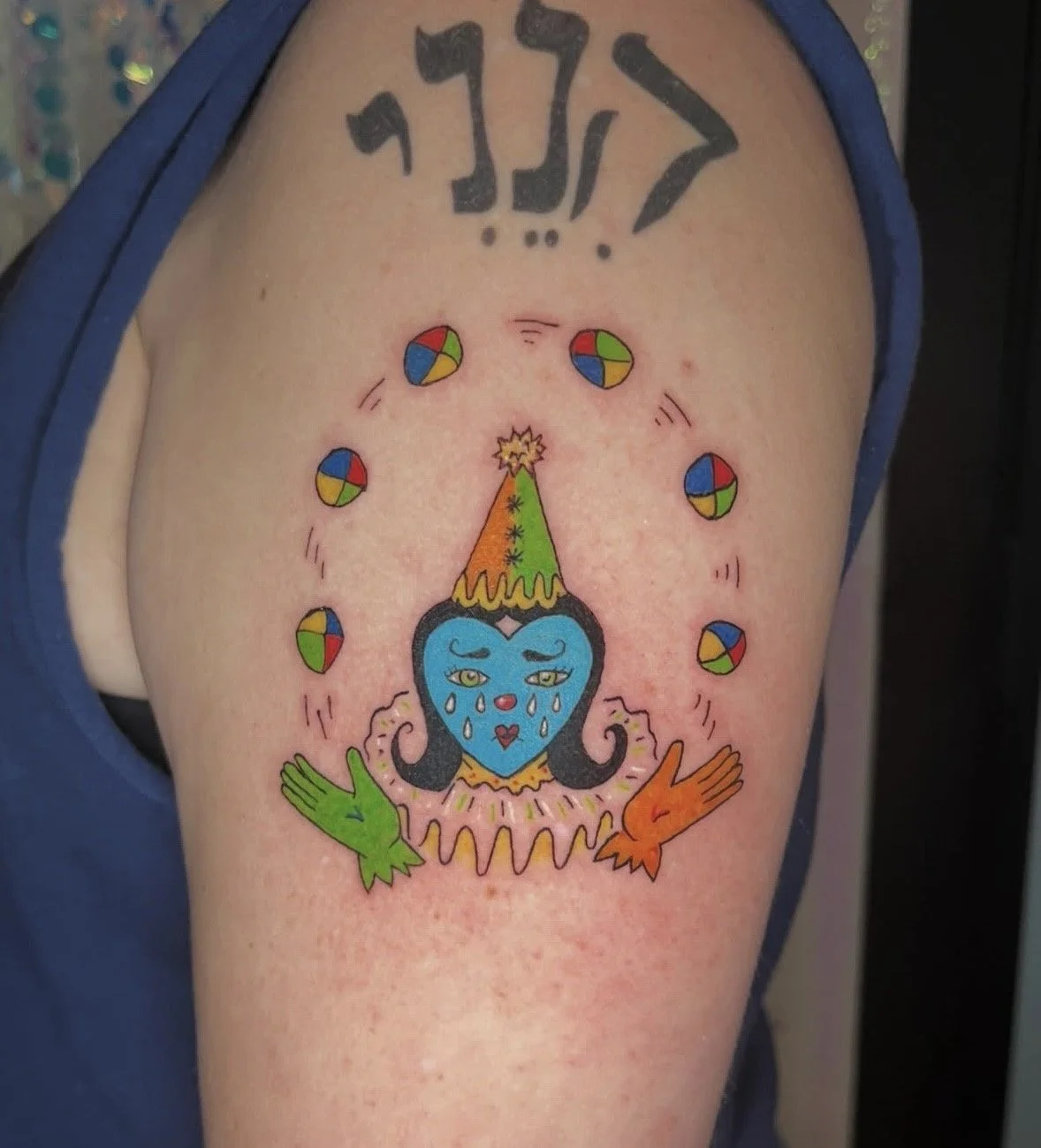 Colorful tattoo on upper arm of a clown with a blue face, tears, and a party hat surrounded by small multicolored circles with motion lines. The word 'JFLY' appears above in black gothic letters.