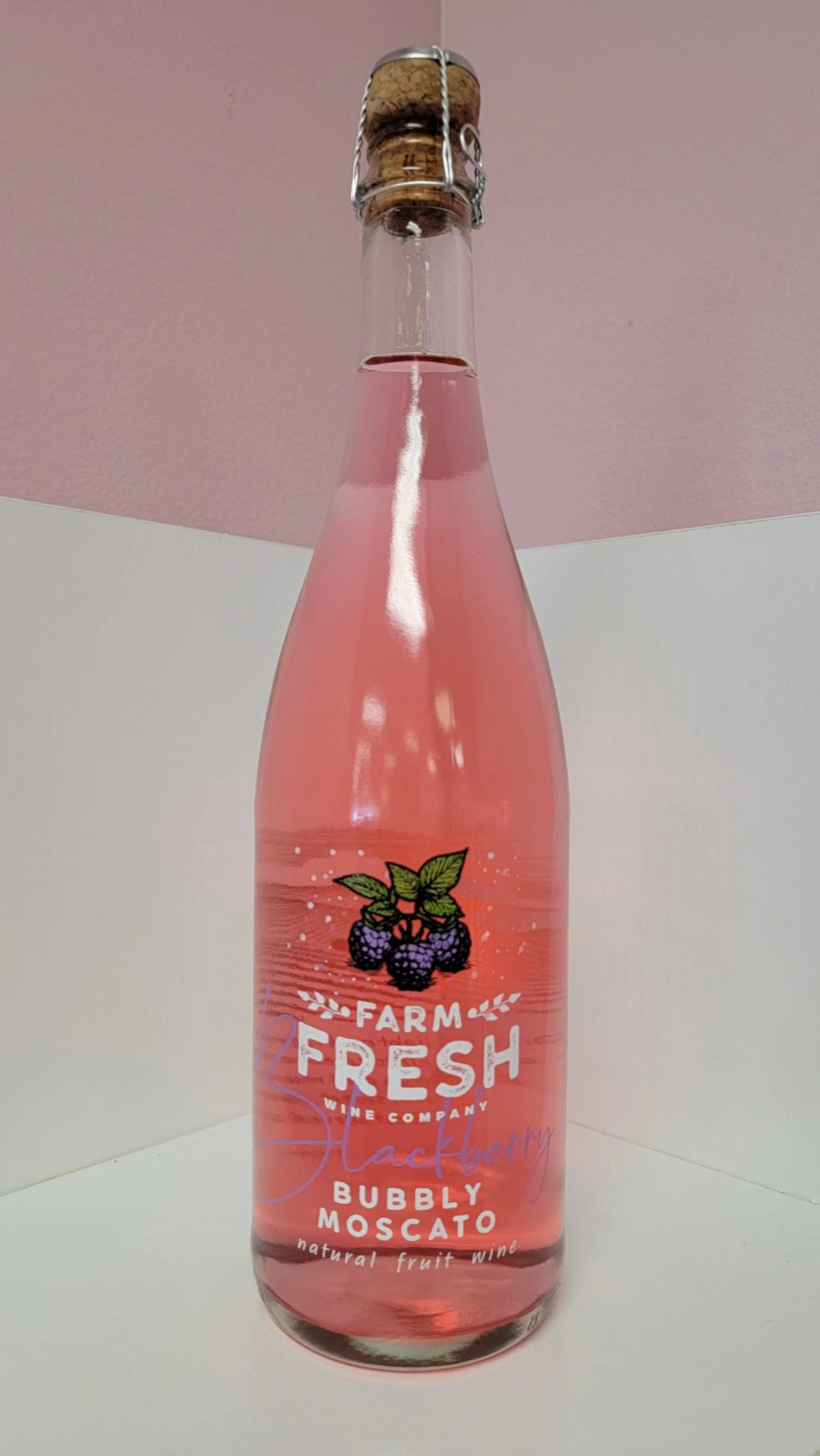 A clear glass bottle of pink raspberry flavored sparkling wine, labeled 'Farm Fresh Raspberry Bubbly Moscato' with a cork and wire cage top.