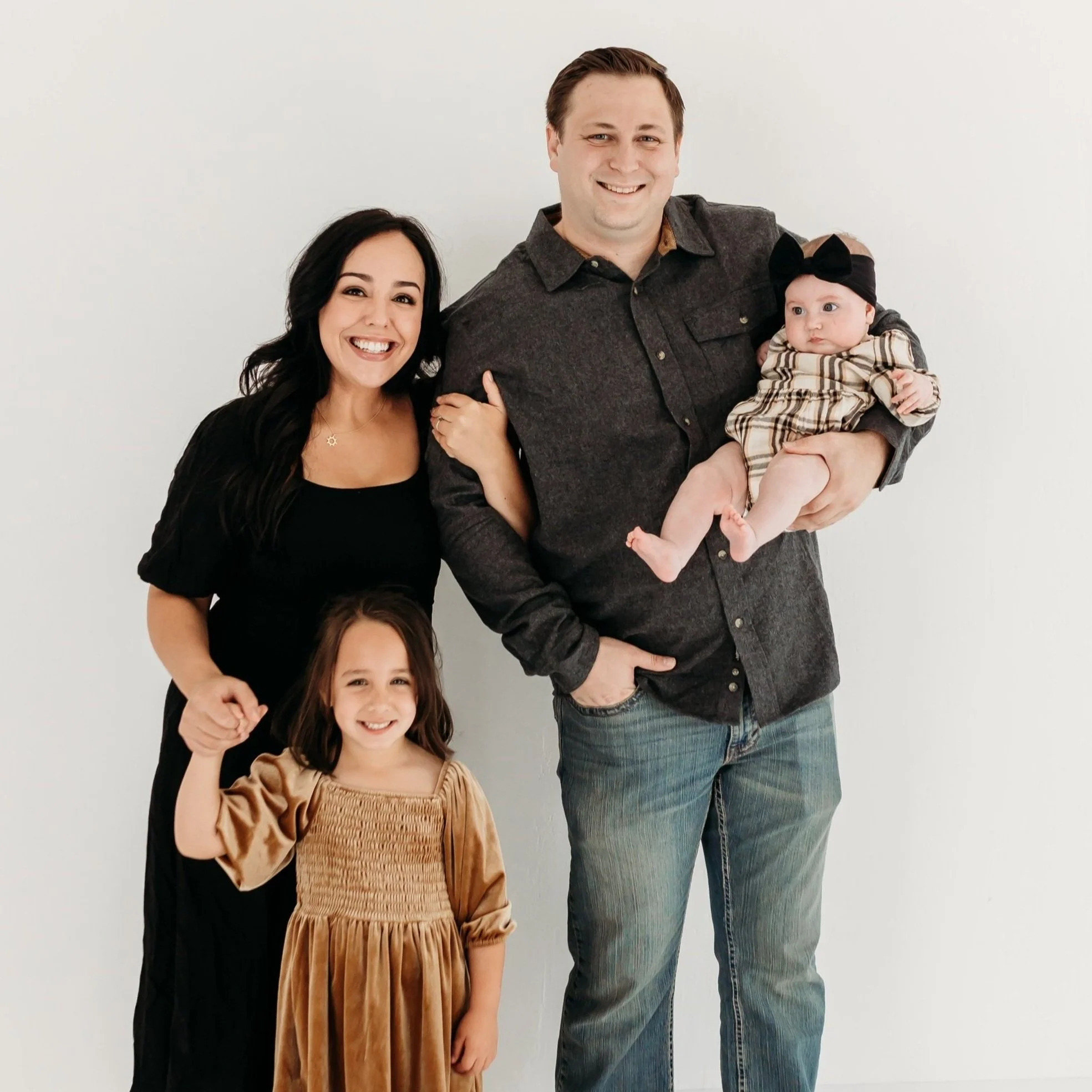 A happy family of four posing for a photo against a plain white wall. The father holds a baby girl, and a woman and a young girl stand beside him, smiling.