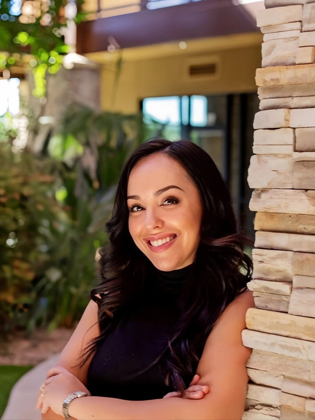 A woman with dark hair and a black sleeveless top smiling while leaning against a stone wall in an outdoor setting.